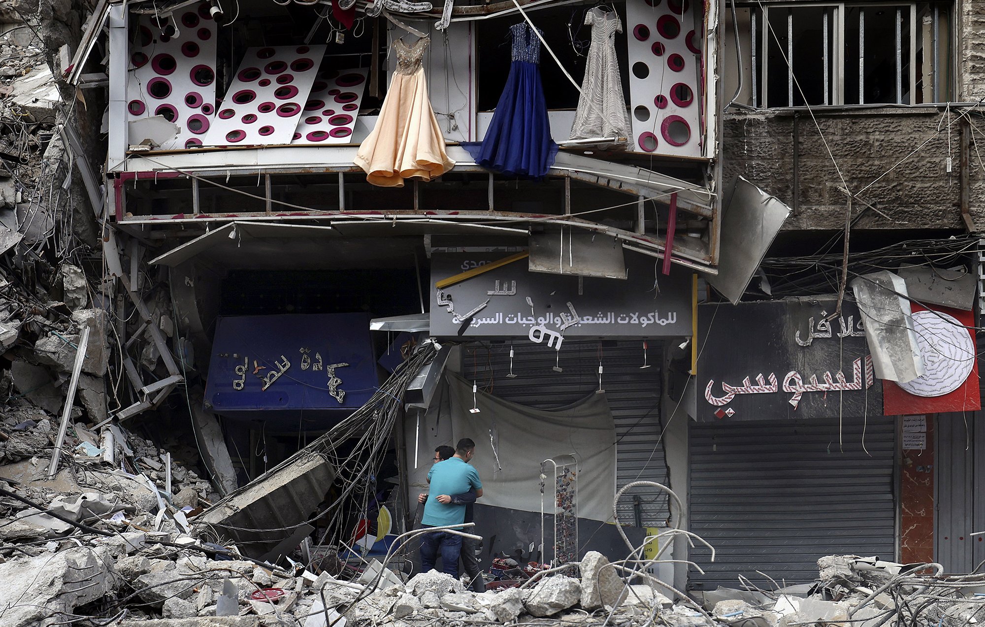L'image montre des débris d'un bâtiment endommagé, probablement en raison d'un conflit ou d'une catastrophe. À l'avant, on peut voir des robes suspendues à l'extérieur d'une boutique, tandis qu'en bas, deux personnes semblent fouiller ou inspecter les lieux. Les murs sont en ruine, et l'environnement dégage une impression de destruction et de désolation. Des enseignes de magasins sont visibles, mais partiellement masquées par les débris.