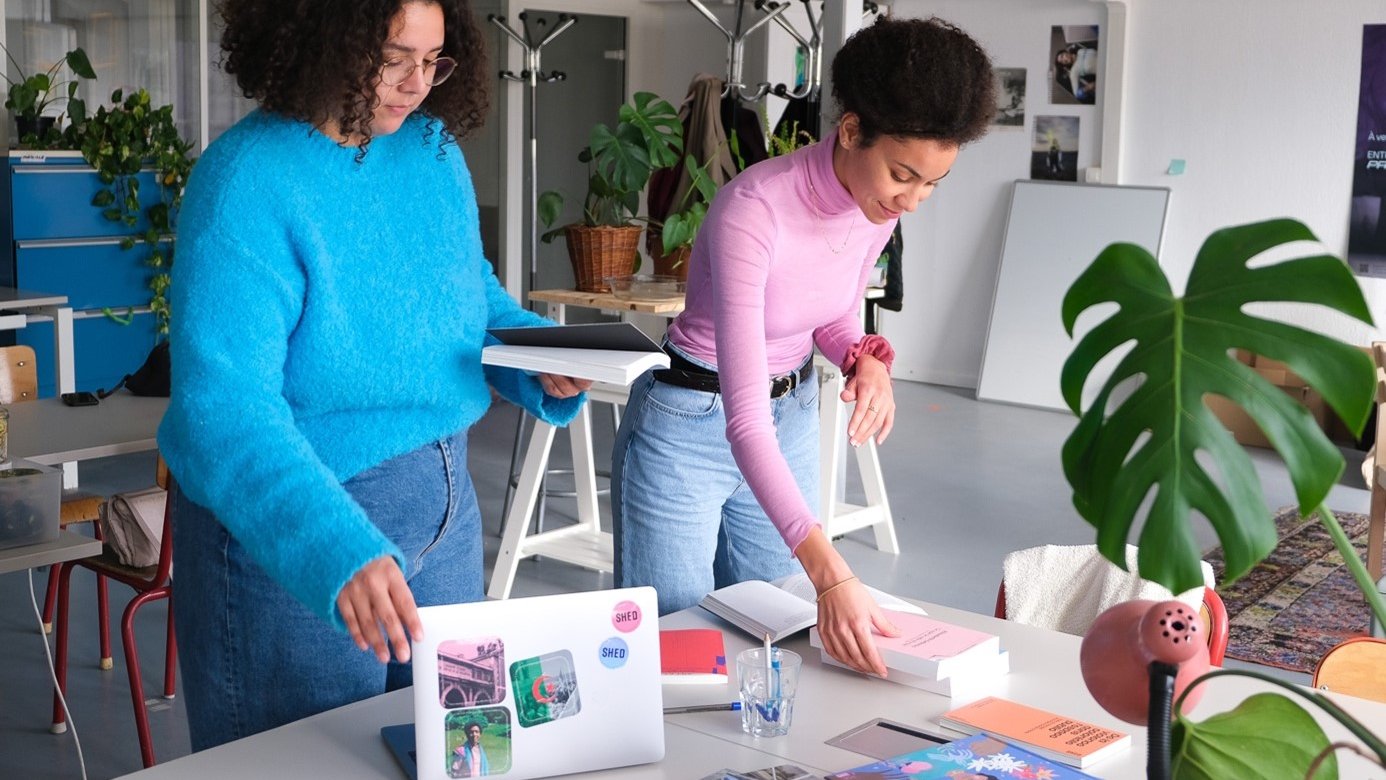 The image shows two women in a modern workspace, engaged in an activity together. One woman, wearing a bright blue sweater, is standing and holding a stack of papers or a folder. The other woman, dressed in a pink top, is bending over a table, arranging or placing items. The table is cluttered with books, a laptop, and other materials, indicating a creative environment. There are plants in the background, adding a touch of greenery to the space. The overall atmosphere appears collaborative and productive.