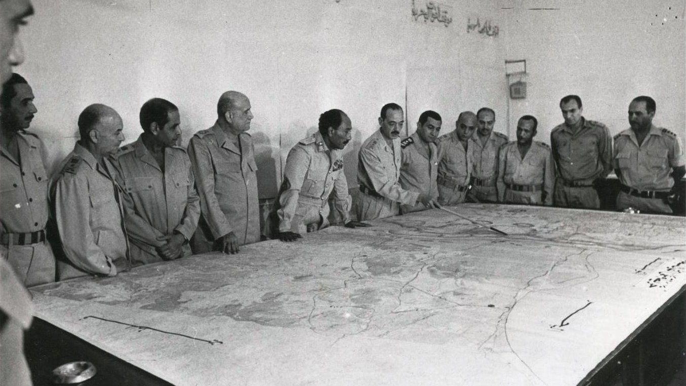 L'image montre un groupe d'hommes en uniformes militaires se tenant autour d'une grande carte posée sur une table. Ils semblent concentrés et discutent de la carte, qui est détaillée avec des lignes et des zones géographiques. L'environnement est clair et sobre, avec des murs nus. L'atmosphère semble sérieuse, probablement en rapport avec des stratégies militaires.