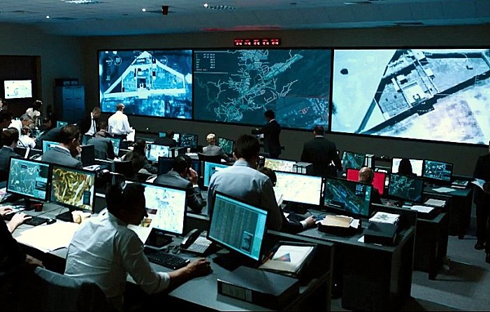 L'image montre une salle de commandes avec de nombreuses personnes travaillant devant des ordinateurs. Il y a plusieurs écrans géants affichant des cartes et des informations, et l'atmosphère semble intense et concentrée. Les employés, habillés en costumes ou en tenues professionnelles, semblent analyser des données ou surveiller des opérations en temps réel. La pièce est éclairée par la lumière des écrans, créant une ambiance technologique et dynamique.