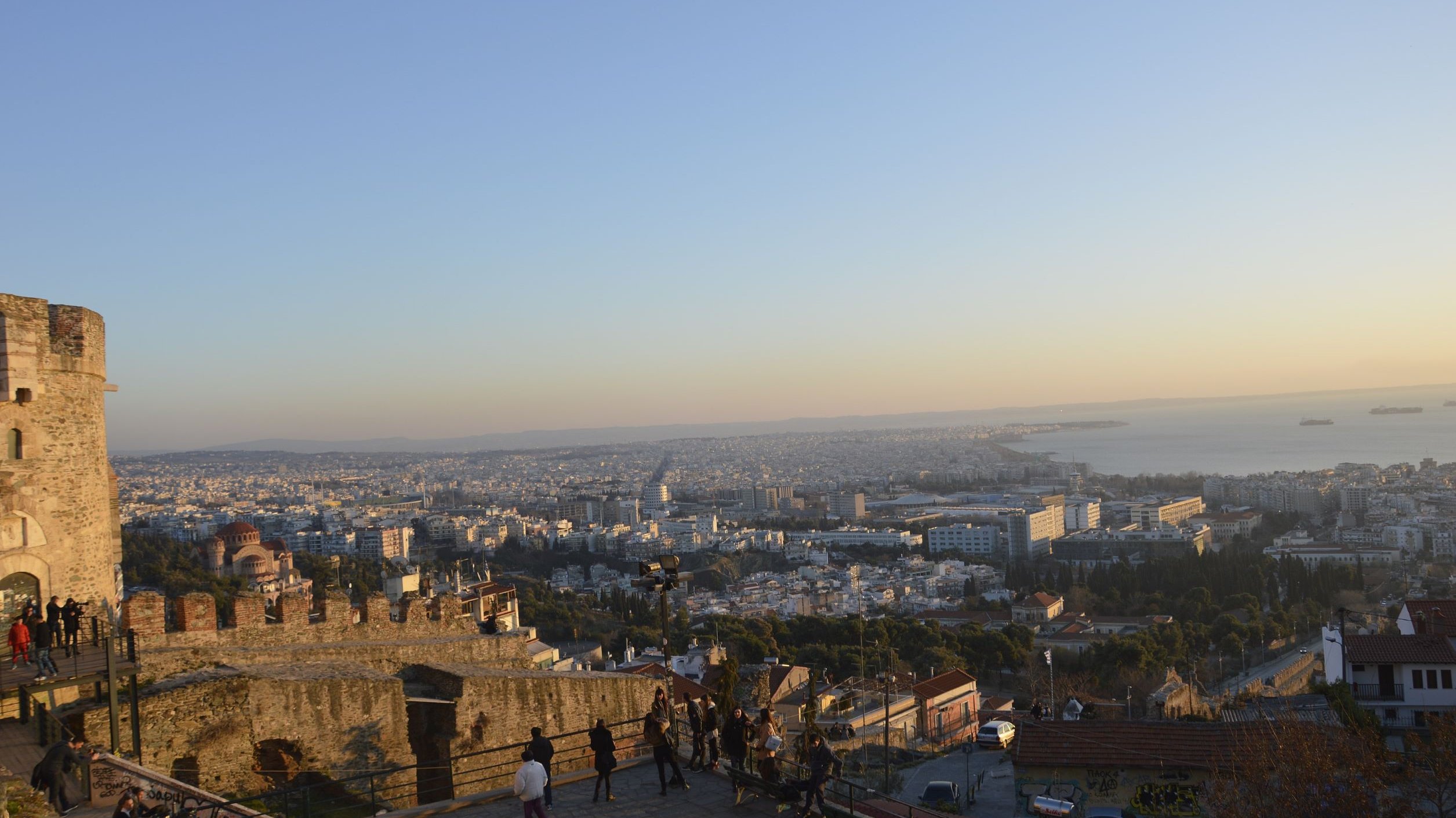 L'image montre une vue panoramique d'une ville au crépuscule. On peut voir un paysage urbain avec de nombreux bâtiments s'étendant jusqu'à l'horizon. À droite, la mer s'étend sur plusieurs kilomètres, et on peut apercevoir quelques bateaux au loin. Au premier plan, on distingue une forteresse ou un château en ruine, avec des murs en pierre. Des personnes se promènent le long d'un chemin, profitant de la vue. La lumière du soir donne une ambiance chaleureuse à la scène.