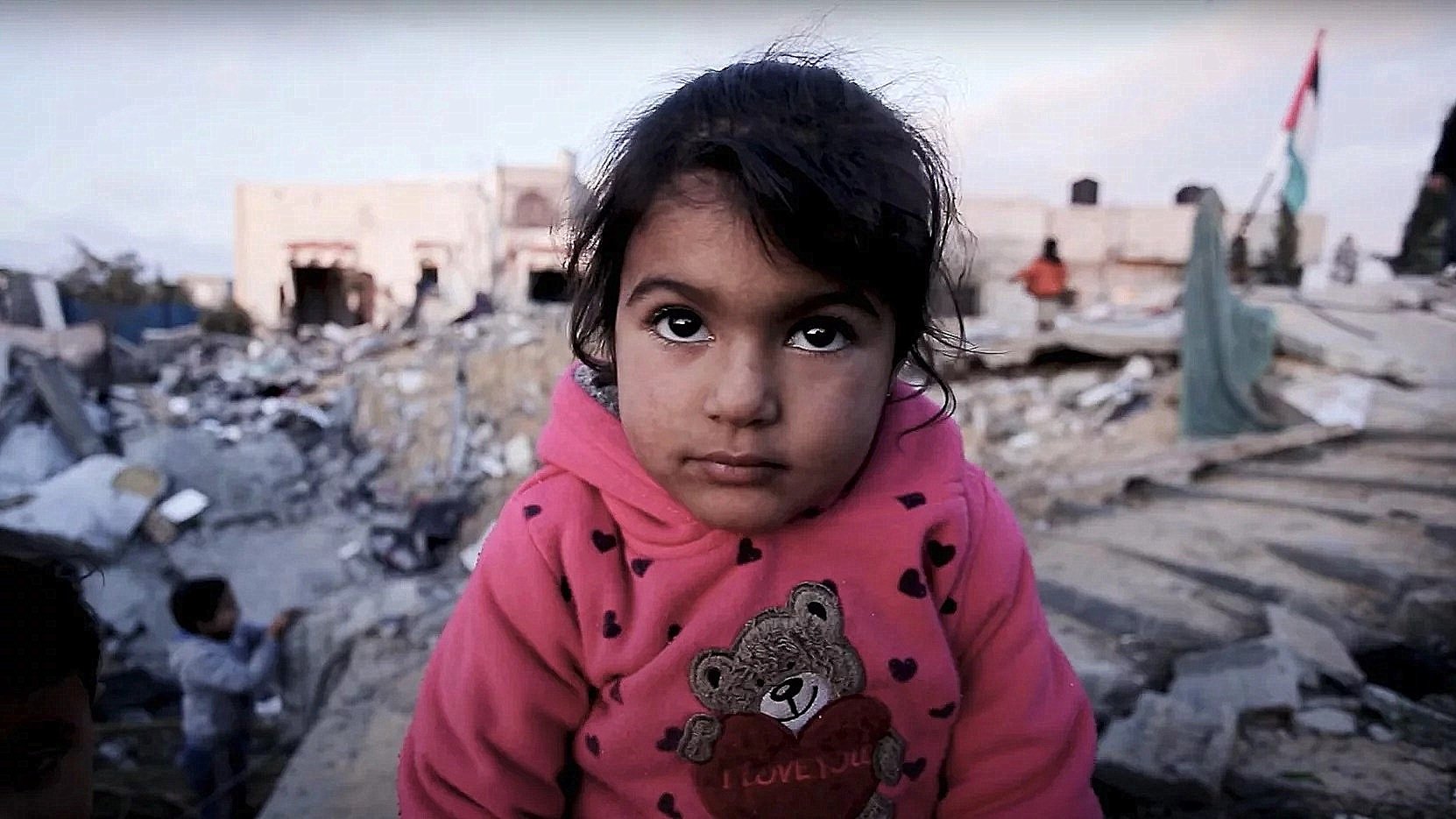 L'image montre une petite fille avec un regard expressif, vêtue d'un sweat à capuche rose orné d'un ours. Elle se trouve dans un environnement dévasté, entourée de décombres et de bâtiments en ruines. En arrière-plan, on aperçoit d'autres enfants. L'atmosphère semble chargée d'une grande tristesse et d'une vulnérabilité palpable.
