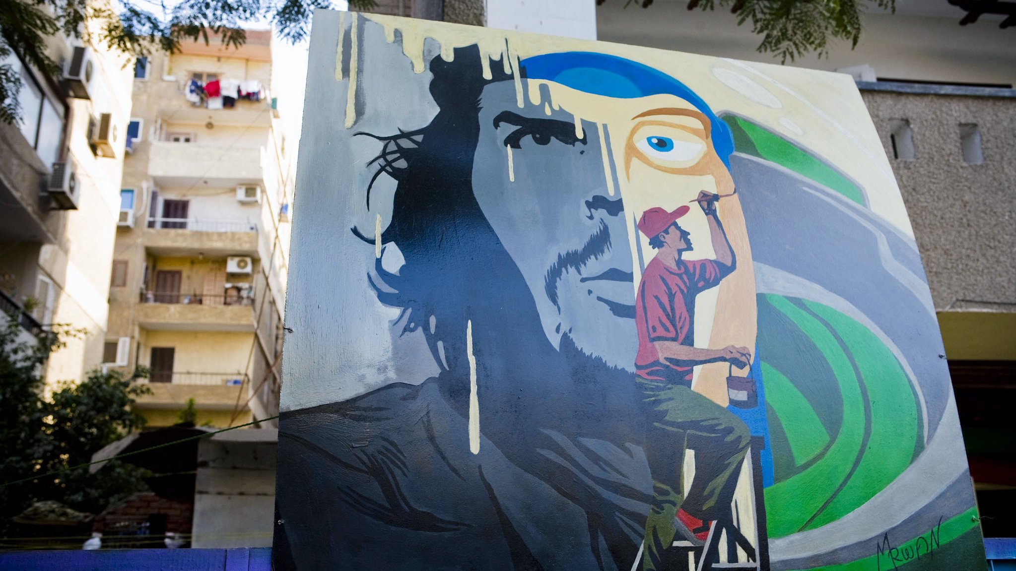 تظهر في الصورة لوحة فنية كبيرة على حائط، تمثل وجه رجل بشكل جزئي، حيث يتواجد شخص آخر على سُلّم يرسم تفاصيل إضافية. الخلفية تحتوي على ألوان زاهية، مع تجسيد ذوات رموز فنية. كما يُمكن رؤية بعض الشقق السكنية في الخلف بالإضافة إلى الملابس المعلقة على الشرفات. تعكس الصورة أجواء حضرية وفنية.