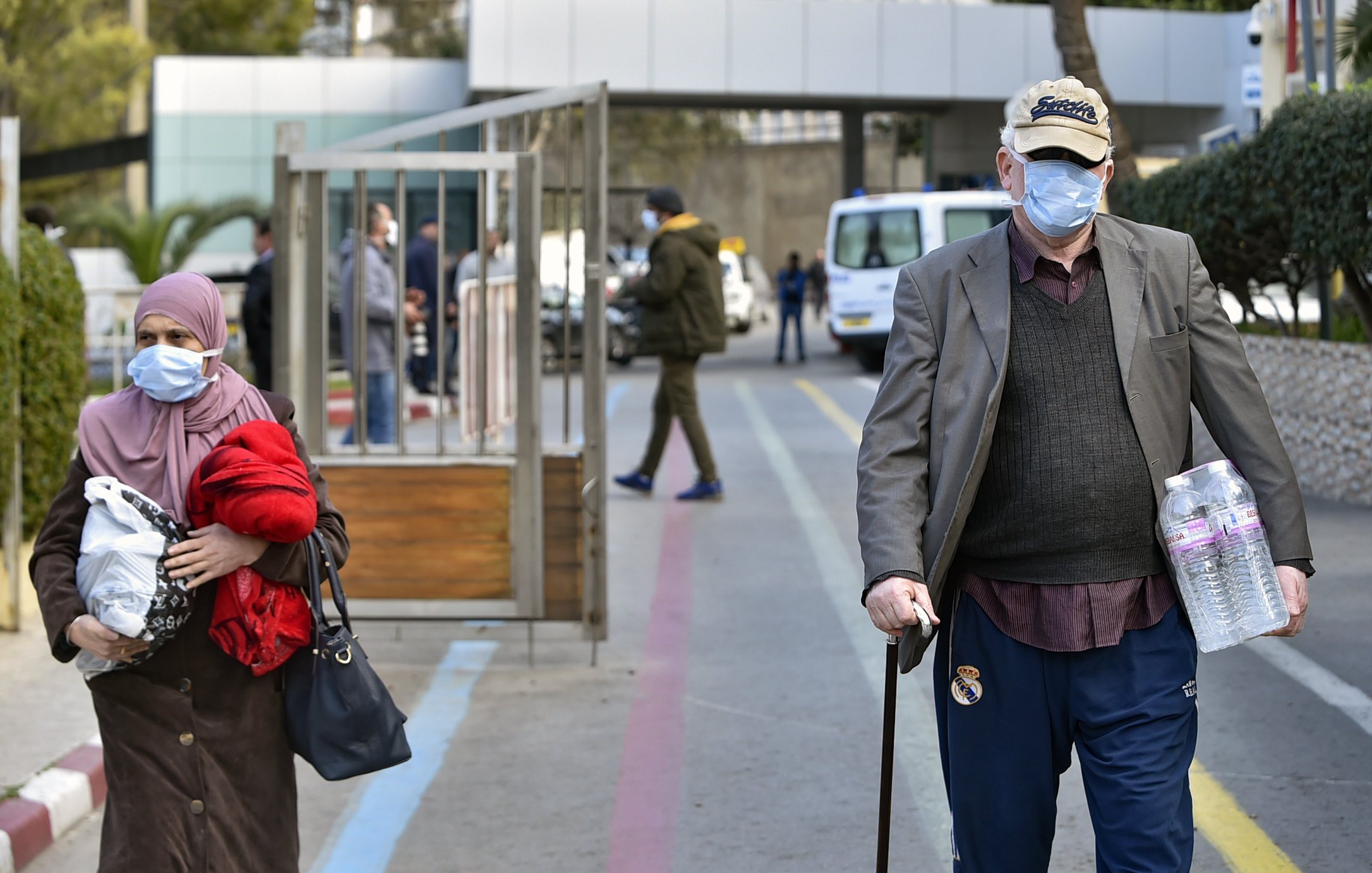 L'image montre une scène urbaine où deux personnes marchent dans une rue. À gauche, une femme portant un foulard et une veste rouge tient un sac, tandis qu'à droite, un homme âgé, avec une canne et un chapeau, marche en portant deux bouteilles d'eau. Les deux portent des masques faciaux, suggérant un contexte sanitaire. En arrière-plan, on aperçoit des véhicules et des bâtiments.