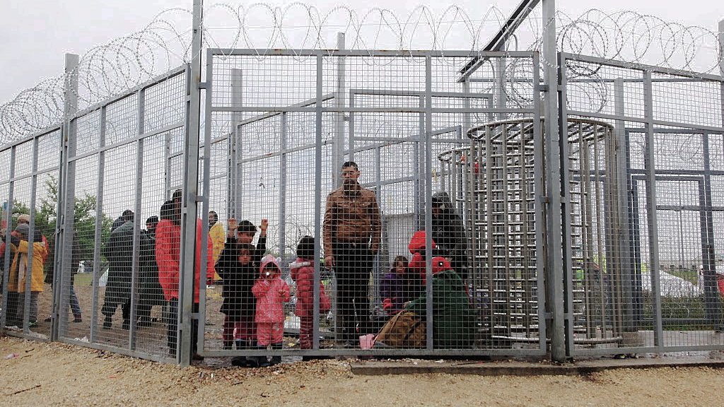 L'image montre un groupe de personnes à l'intérieur d'une structure grillagée, entourée de barbelés. On peut voir des adultes et des enfants, certains portant des vêtements colorés, semblant attendre dans un espace très restreint. L'ambiance semble préoccupante, suggérant une situation de confinement ou d'attente difficile. Le ciel est nuageux, ce qui contribue à une impression de morosité.