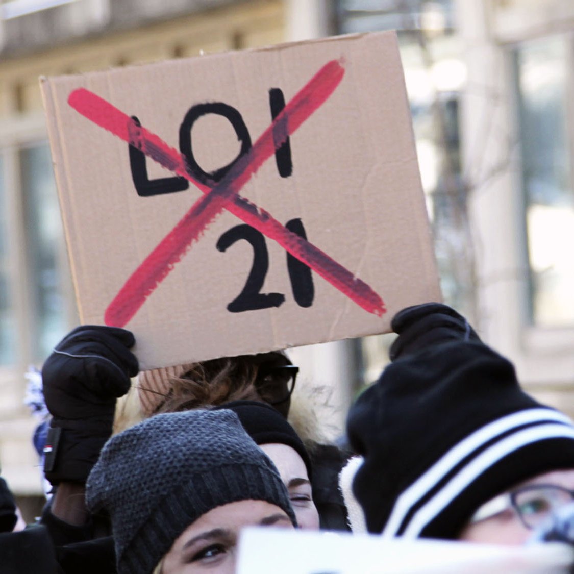 تظهر الصورة مجموعة من الأشخاص في تجمع أو احتجاج، حيث يحمل أحدهم لافتة تشير إلى معارضة لشيء ما. اللافتة مكتوبة عليها "21" مع علامة "إكس" حمراء تدل على رفض هذا الرقم أو القانون. يبدو أن المشاركين يرتدون ملابس شتوية، مما يوحي أن الحدث وقع في فصل الشتاء. تعكس الصورة روح الحماس والمشاركة من قبل المحتجين.