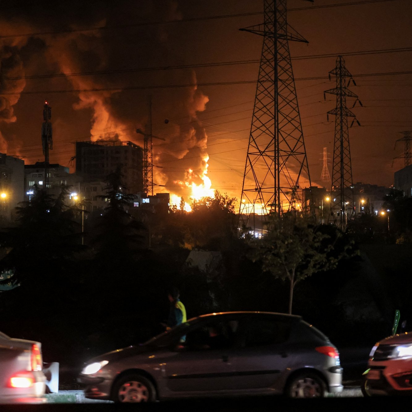 Flammes illuminent la nuit, voitures passent près d'une scène de chaos urbain.