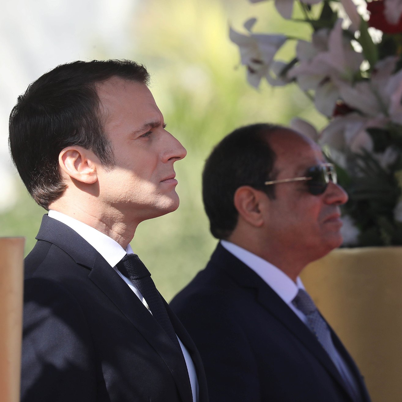 الصورة تظهر رجلين standing بجانب بعضهما البعض. الرجل على اليسار يرتدي بدلة داكنة مع ربطة عنق، بينما الرجل على اليمين يرتدي نظارات شمسية وبدلة رسمية. الخلفية تحتوي على زهور وأشجار، مما يضفي جوًا من الاحتفال أو المناسبة الرسمية. تعكس الصورة موقفًا رسميًا وجديًا بين الشخصين.