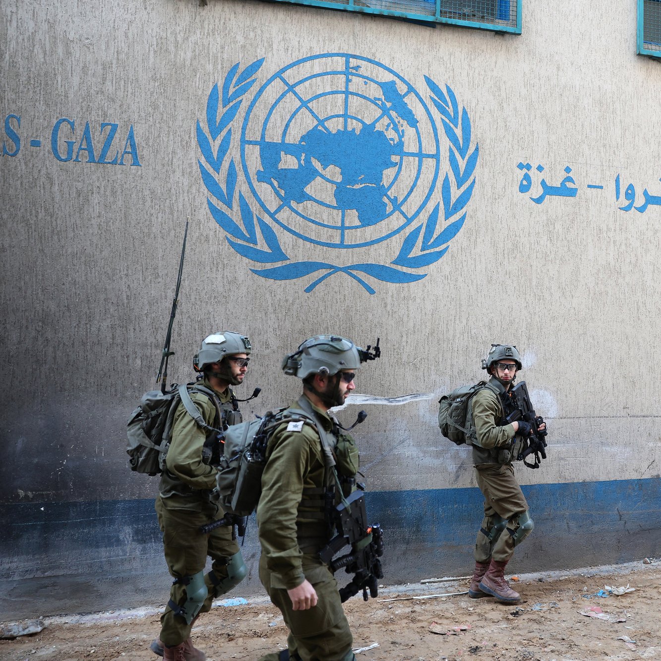L'image montre des soldats en uniforme marchant près d'un bâtiment, probablement associé à l'ONU, avec un logo visible sur le mur. Leurs tenues militaires indiquent qu'ils sont en mission. L'environnement semble urbain, avec des structures typiques d'une zone habitée. L'atmosphère peut être perçue comme tendue en raison de la présence militaire.