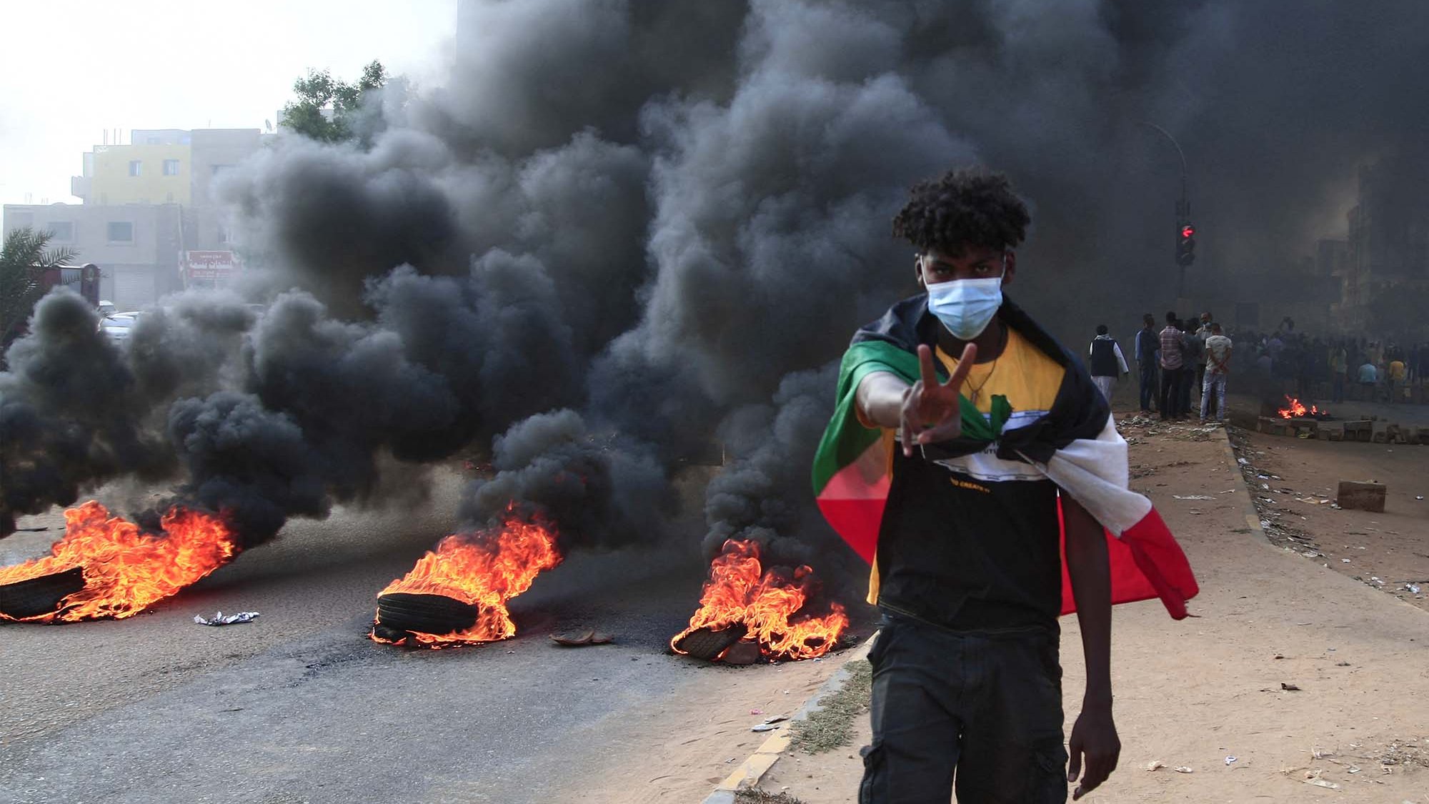 L'immagine mostra un giovane che cammina lungo una strada, con fumi neri che si alzano dietro di lui. Ci sono pneumatici in fiamme che bruciano ai lati della strada. Il ragazzo indossa una mascherina e fa un gesto di pace con la mano. Sullo sfondo, si notano altre persone che osservano la scena. L'atmosfera sembra intensa e carica di tensione.