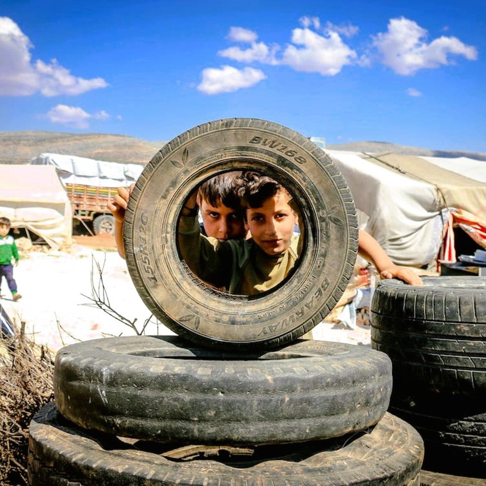 L'image montre deux enfants regardant à travers un pneu, dans un environnement qui semble être un camp de réfugiés. On peut voir des pneus empilés autour d'eux et des abris simples en arrière-plan. Le ciel est dégagé avec quelques nuages. Les enfants affichent une expression curieuse, et la scène dépeint un moment de jeu au milieu d'un cadre difficile.