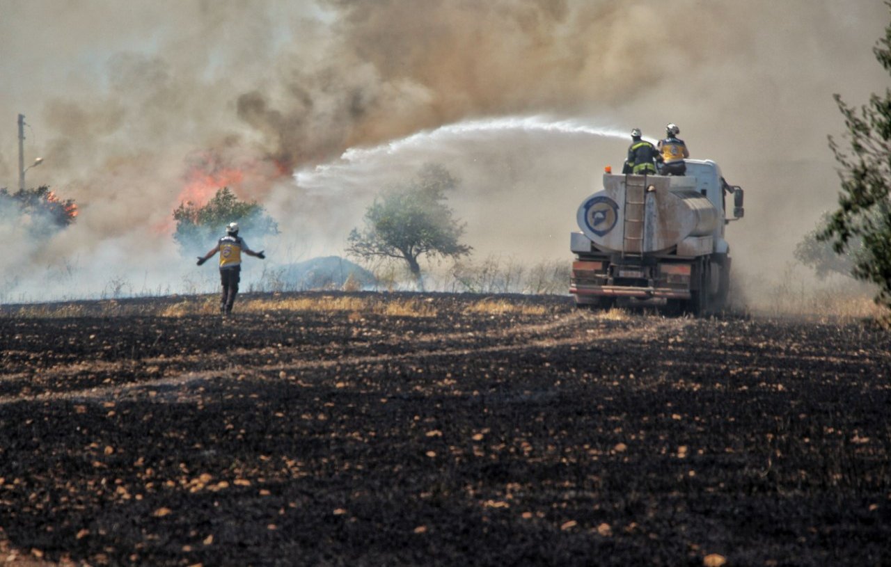 L'image montre une scène de lutte contre un incendie. On voit des pompiers en action, utilisant un camion-citerne pour éteindre les flammes qui s'élèvent dans la fumée. Au premier plan, un pompier semble se diriger vers le feu, tandis que l'arrière-plan montre un paysage noirci, témoignant des dégâts causés par l'incendie. Les arbres sont entourés de fumée, créant une atmosphère de tension et d'urgence.