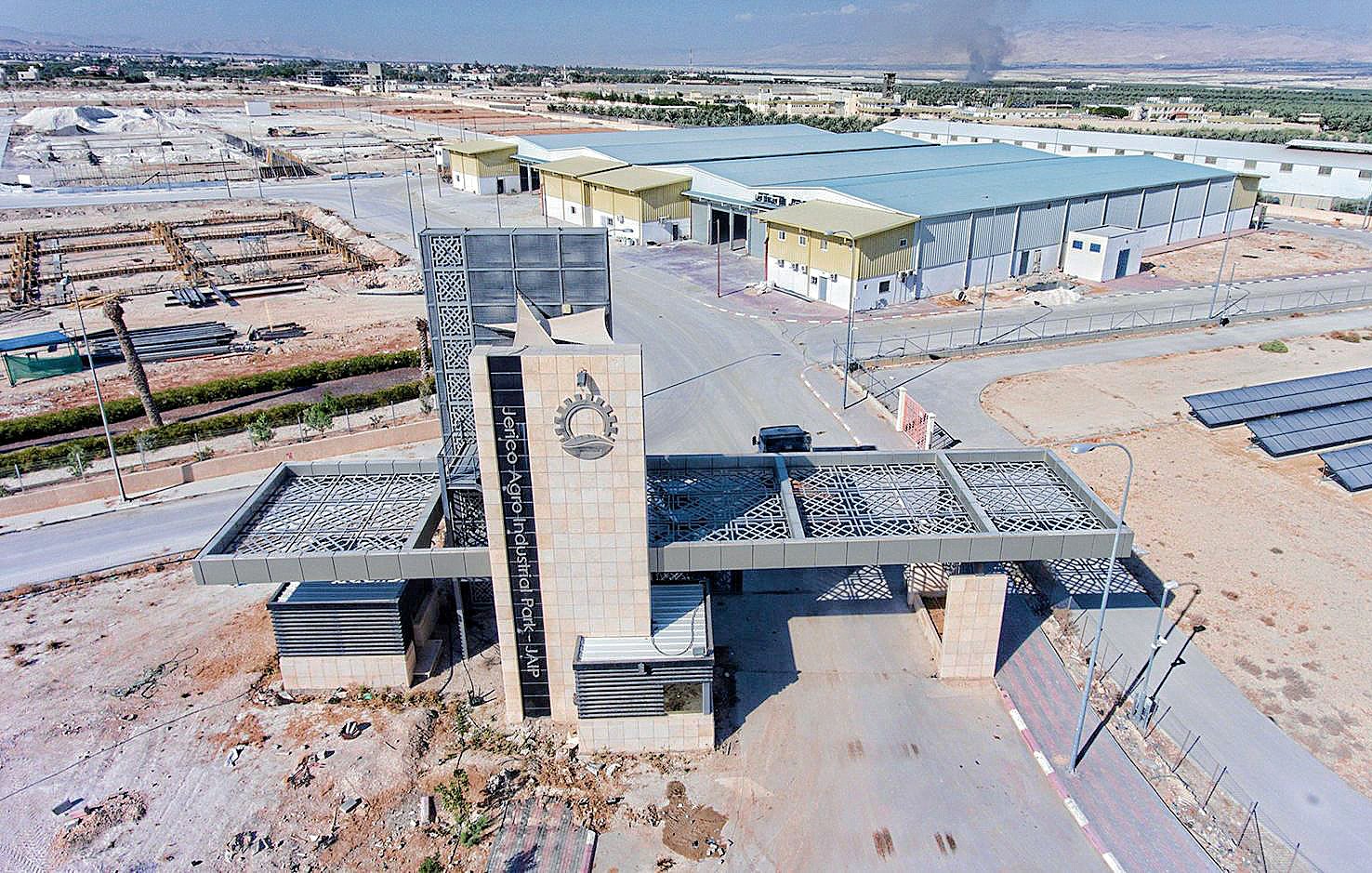 L'image montre un complexe industriel ou commercial avec une entrée bien marquée. On peut voir un portail moderne, probablement en métal et en pierre, qui semble être la pièce maîtresse de l'entrée. À l'arrière plan, il y a plusieurs bâtiments de différentes tailles, dont un plus grand qui pourrait servir de site de production ou d'entrepôt. Le terrain environnant est désertique,avec quelques infrastructures comme des panneaux solaires. En arrière-plan, on distingue un nuage de fumée, suggérant une activité ou un incendie à proximité.