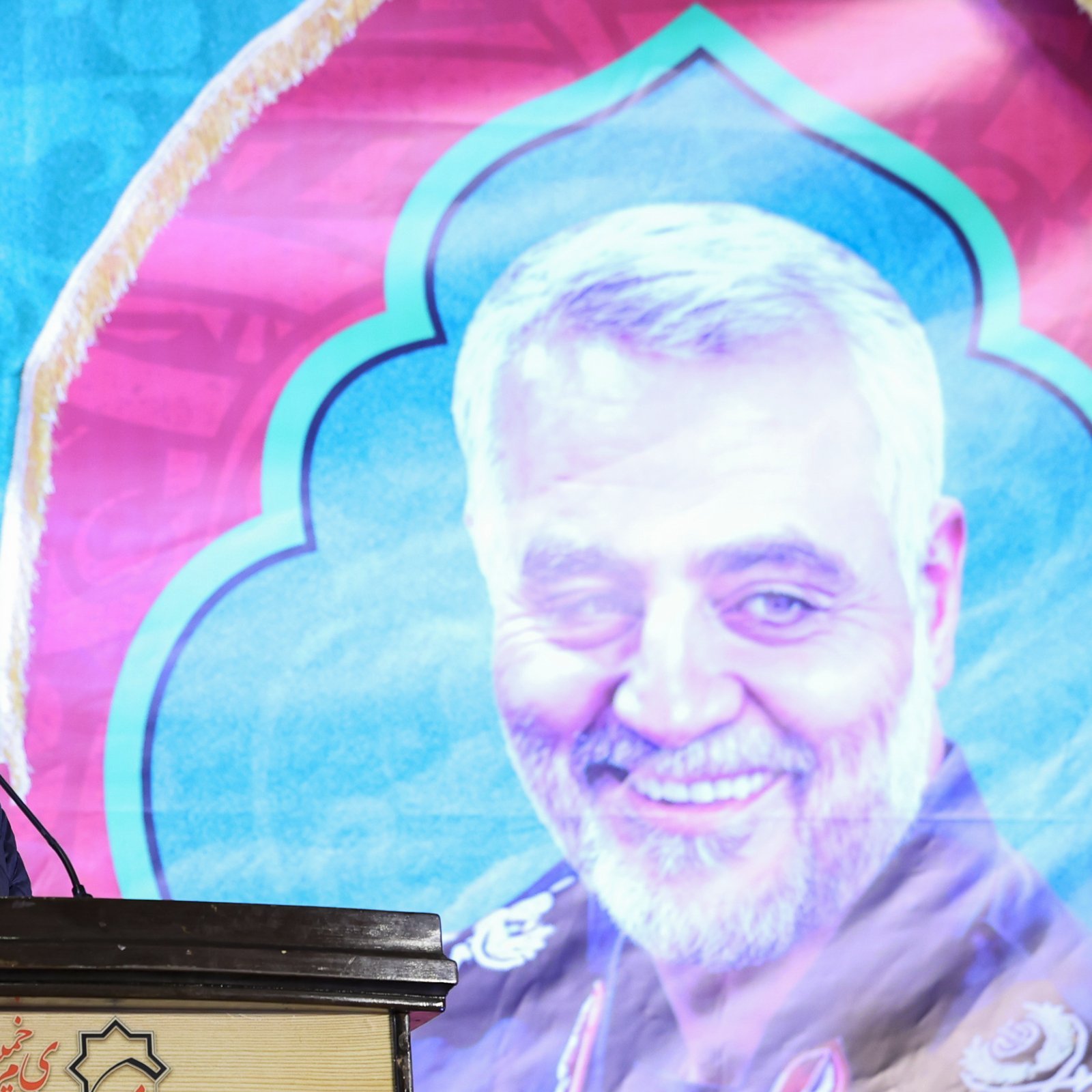 در این تصویر، شخصی در حال سخنرانی در یک مراسم دیده می‌شود. او پشت میکروفون قرار دارد و به طرف جمعیت صحبت می‌کند. در پس‌زمینه، تصویری بزرگ از فرد دیگری که لباس نظامی به تن دارد، به‌وضوح قابل مشاهده است. طراحی پس‌زمینه رنگی و با الگوهای خاص است که به مراسم زیبایی می‌بخشد. فضای کلی مراسم جدی و متعهد به هدف خاصی به نظر می‌رسد.