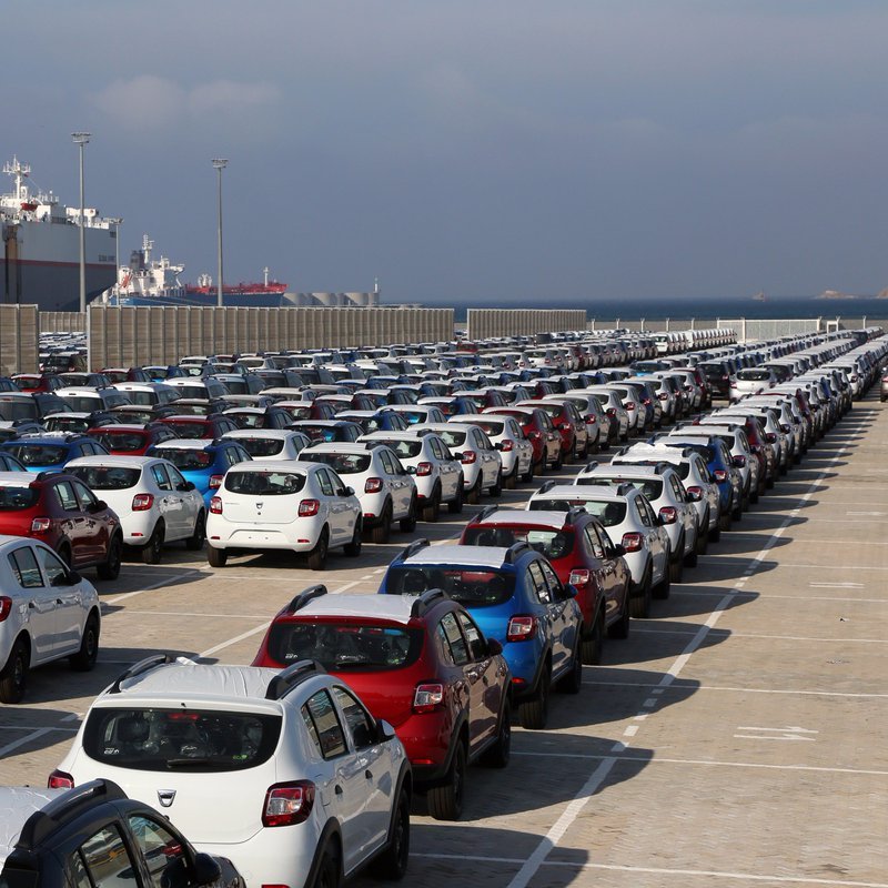 تظهر الصورة مشهداً لموقف سيارات كبير في ميناء، حيث تكون السيارات مصطفة بشكل منظم. يوجد مجموعة متنوعة من السيارات بألوان مختلفة، مثل الأبيض والأزرق والأحمر. في الخلفية، يمكن رؤية سفن راسية في الميناء تحت سماء زرقاء مع بعض السحب. يبدو أن المكان مخصص لتخزين السيارات، مما يوحي بأنه يعتمد على عمليات الشحن والنقل.