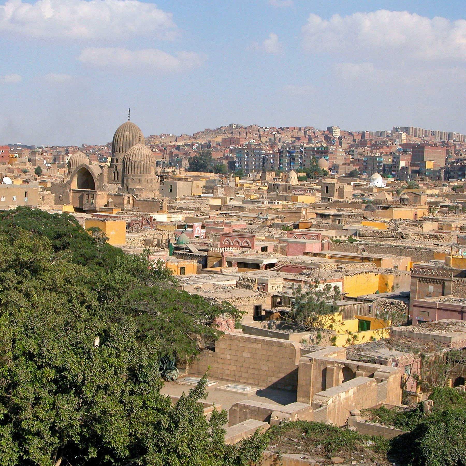 L'image montre une vue panoramique d'une ville historique, probablement un quartier ancien avec des bâtiments en pierre. On peut voir plusieurs coupoles et minarets qui témoignent de l'architecture traditionnelle. Les toits sont variés, certains sont plats et d'autres sont plus élevés, tandis que la végétation semble présente avec des arbres. En arrière-plan, on aperçoit des collines et d'autres bâtiments, suggérant une ville vaste et densément peuplée. Le ciel est clair avec quelques nuages, donnant une atmosphère lumineuse à la scène.