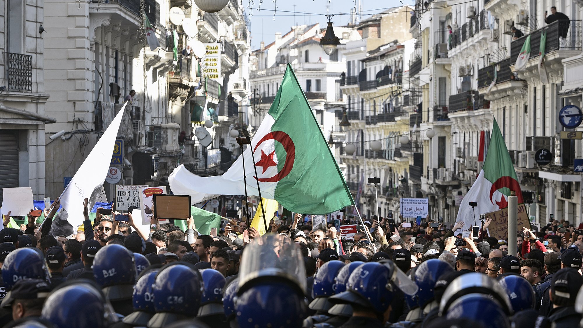 L'image montre une foule rassemblée dans une rue animée, probablement dans une ville algérienne, en période de manifestation. Des personnes portent des drapeaux algériens et tiennent des pancartes, tandis qu'un groupe de policiers en uniforme, portant des casques, se tient en ligne devant eux. Les bâtiments alentours sont typiques d'une architecture urbaine, ornés de décorations. L'atmosphère semble être celle d'une démonstration populaire, avec un fort engagement civique visible parmi les manifestants.