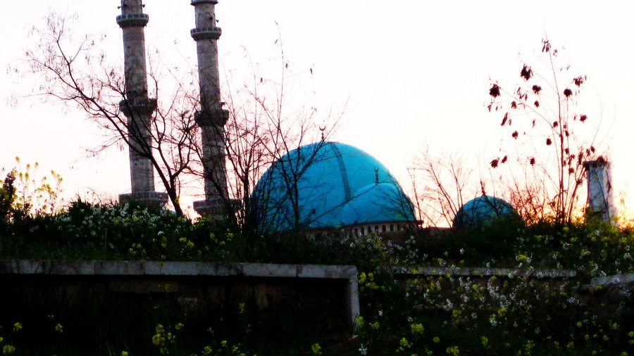 L'image montre un paysage au coucher du soleil avec deux minarets élancés se dressant vers le ciel. En arrière-plan, on peut apercevoir un grand dôme turquoise, probablement d'une mosquée, entouré de végétation. Des fleurs sauvages poussent au premier plan, ajoutant une touche de couleur et de vie à la scène. Les teintes douces du coucher de soleil créent une atmosphère paisible et tranquille.