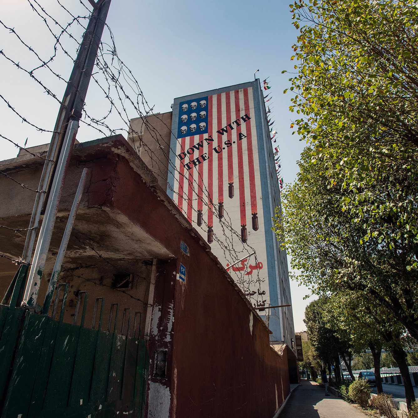 L'image montre un mur d'un bâtiment avec une grande peinture représentant un drapeau américain à rayures. Le mur est bordé par une clôture en barbelés, et on peut voir des arbres sur le côté. Le paysage est urbain, avec un chemin pavé qui suggère une rue à proximité. La composition évoque un contraste entre l'élément décoratif du drapeau et la présence de la clôture de sécurité.