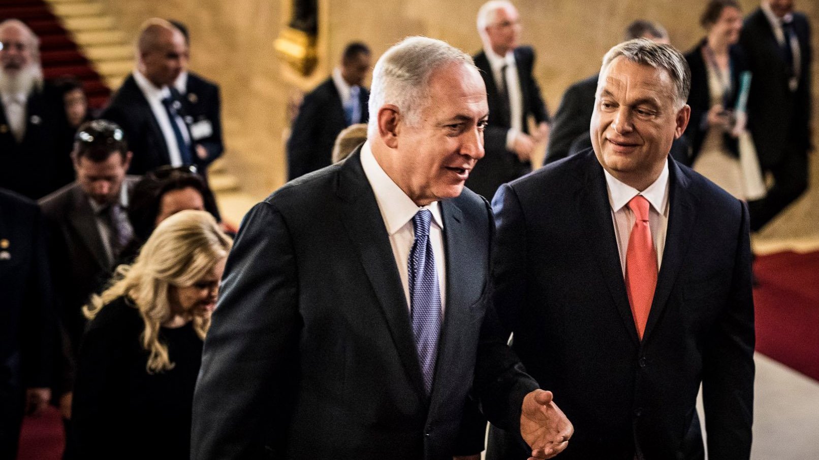 در این تصویر، دو مرد در حال صحبت و قدم زدن به سمت جلو هستند. یکی از آنها لباس سیاه و کراوات آبی به تن دارد و دیگری لباس رسمی با کراوات صورتی بر تن دارد. تعدادی از افراد دیگر نیز در پس‌زمینه وجود دارند که به نظر می‌رسد در یک مکان رسمی یا مراسم حضور دارند. فضای تصویر نشان‌دهنده یک موقعیت مهم یا رسمی است.