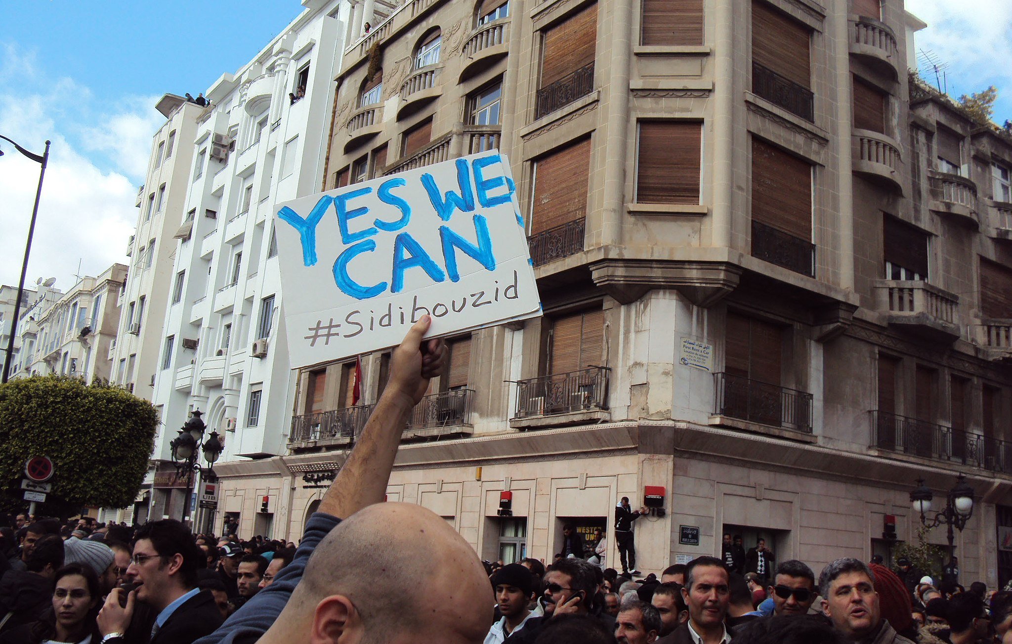 در این تصویر، گروهی از مردم در یک تجمع یا تظاهرات شرکت کرده‌اند. شخصی در جلوی تصویر، تابلویی با نوشته «YES WE CAN» و هشتگ «#Sidibouzid» را در دست دارد. ساختمان‌هایی با معماری مشخص در پس‌زمینه دیده می‌شود و جمعیت به‌نظر معترض یا فعال است. فضای تصویر حالت اجتماعی و سیاسی به خود گرفته و نشان‌دهندهٔ یک حرکت جمعی است.