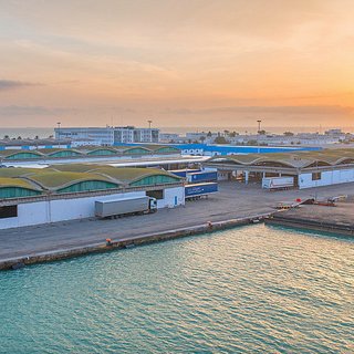 L'image montre un port lors d'un coucher de soleil, avec un paysage industriel. On peut voir des entrepôts aux toits arrondis, bordés par l'eau. Des véhicules et des conteneurs sont visibles, indiquant une activité portuaire. Le ciel est teinté de couleurs chaudes, créant une atmosphère paisible et accueillante.