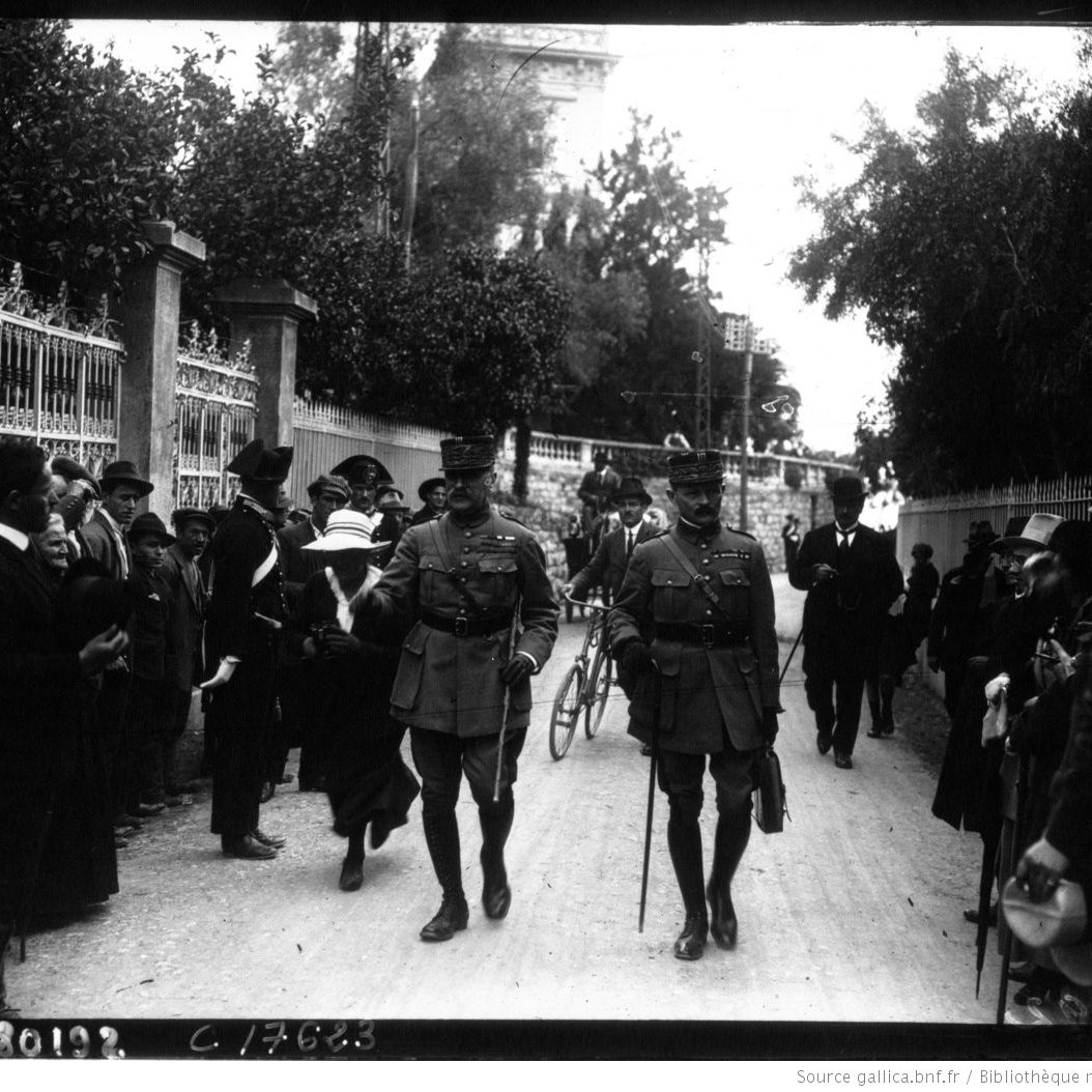 L'image montre une rue entourée de personnes, dont des militaires en uniforme. Deux officiers marchent au premier plan, semblant discuter. On aperçoit des civils sur les côtés, certains regardant vers les officiers. L'ambiance semble solennelle, et l'environnement est arboré, avec des bâtiments en arrière-plan. Les vêtements des personnes indiquent une époque historique. L'image a une qualité noire et blanche, typique des photographies anciennes.