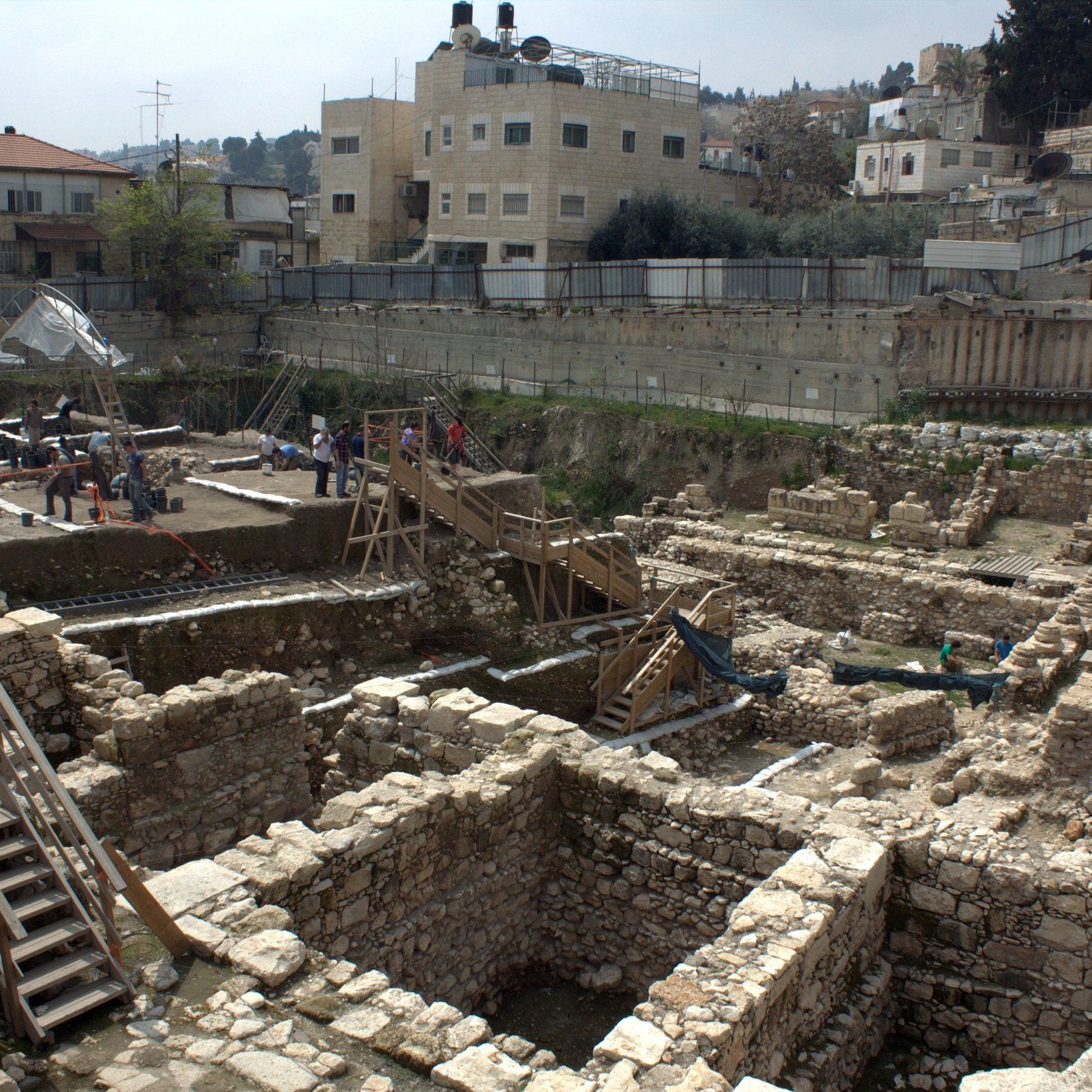 L'image montre des fouilles archéologiques en cours dans une zone urbaine. On peut voir des structures en pierre disposées de manière organisée, avec des vestiges de murs et de fondations visibles. Des scaffolds et des passerelles en bois sont installés pour permettre aux archéologues de travailler et d'accéder facilement aux différentes couches de sol. En arrière-plan, des bâtiments modernes sont présents, ce qui témoigne du mélange entre le patrimoine historique et l'urbanisation contemporaine. Des chercheurs et des travailleurs s'activent autour du site, suggérant une recherche active sur l'histoire de la région.