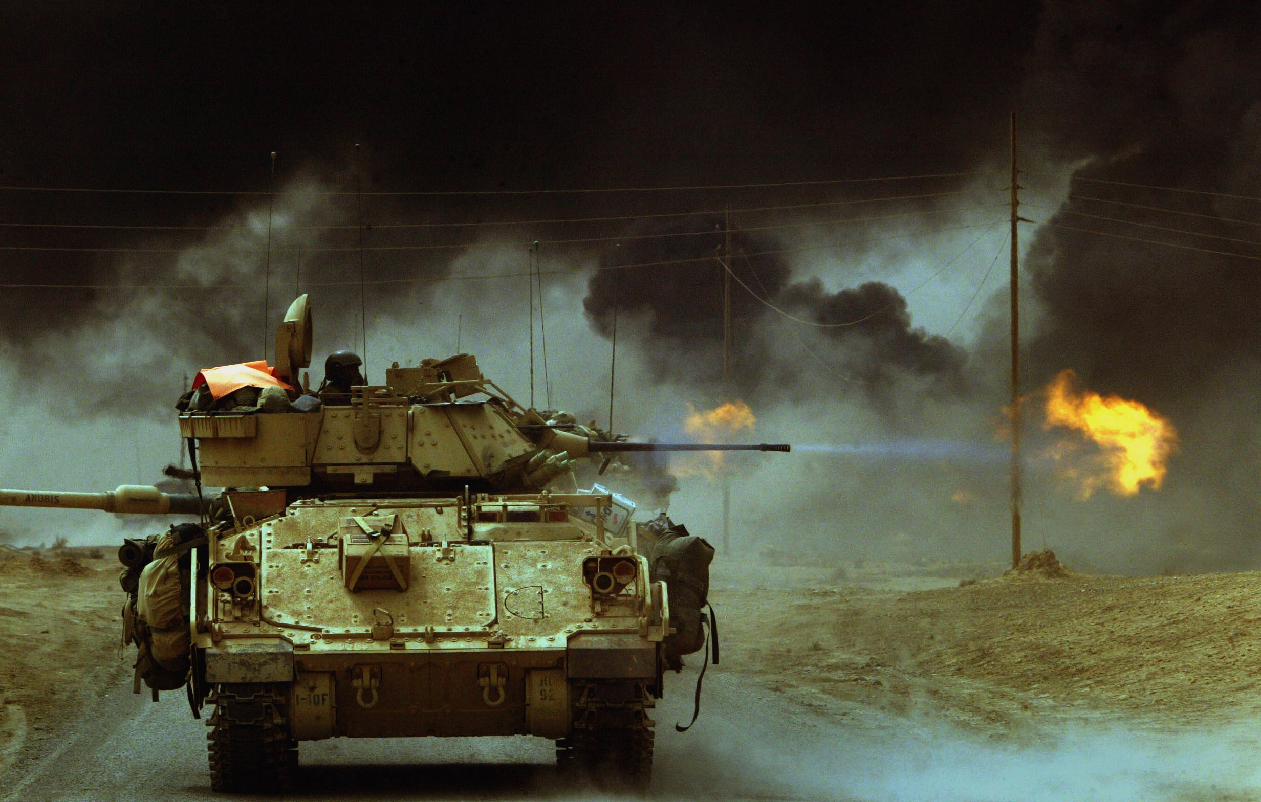 L'image montre un char de combat sur une route, probablement en zone de conflit. Des colonnes de fumée noire s'élèvent vers le ciel, tandis que des flammes jaillissent, suggérant des explosions ou des incendies à proximité. L'ambiance générale est sombre et chaotique, soulignant les effets de la guerre et la destruction présente dans le paysage. Le char est armé et semble se préparer à avancer dans un environnement hostile.