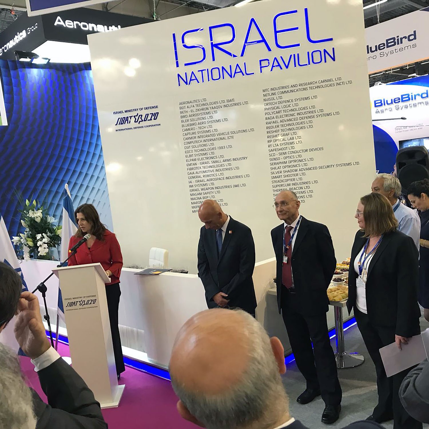 تظهر الصورة مجموعة من الأشخاص في حدث أو معرِض، حيث يتواجد متحدثة أمام منصة تحمل شعار "إسرائيل" وتفاصيل عن الجناح الوطني. هناك بعض الحضور في الخلفية يستمعون بانتباه، بينما يبدو بعضهم منشغلاً بتدوين الملاحظات. المنصة محاطة بالزهور، ويوجد مرافقون يرتدون بدلات رسمية. الأجواء تعكس نشاطًا رسميًا ومهنيًا.