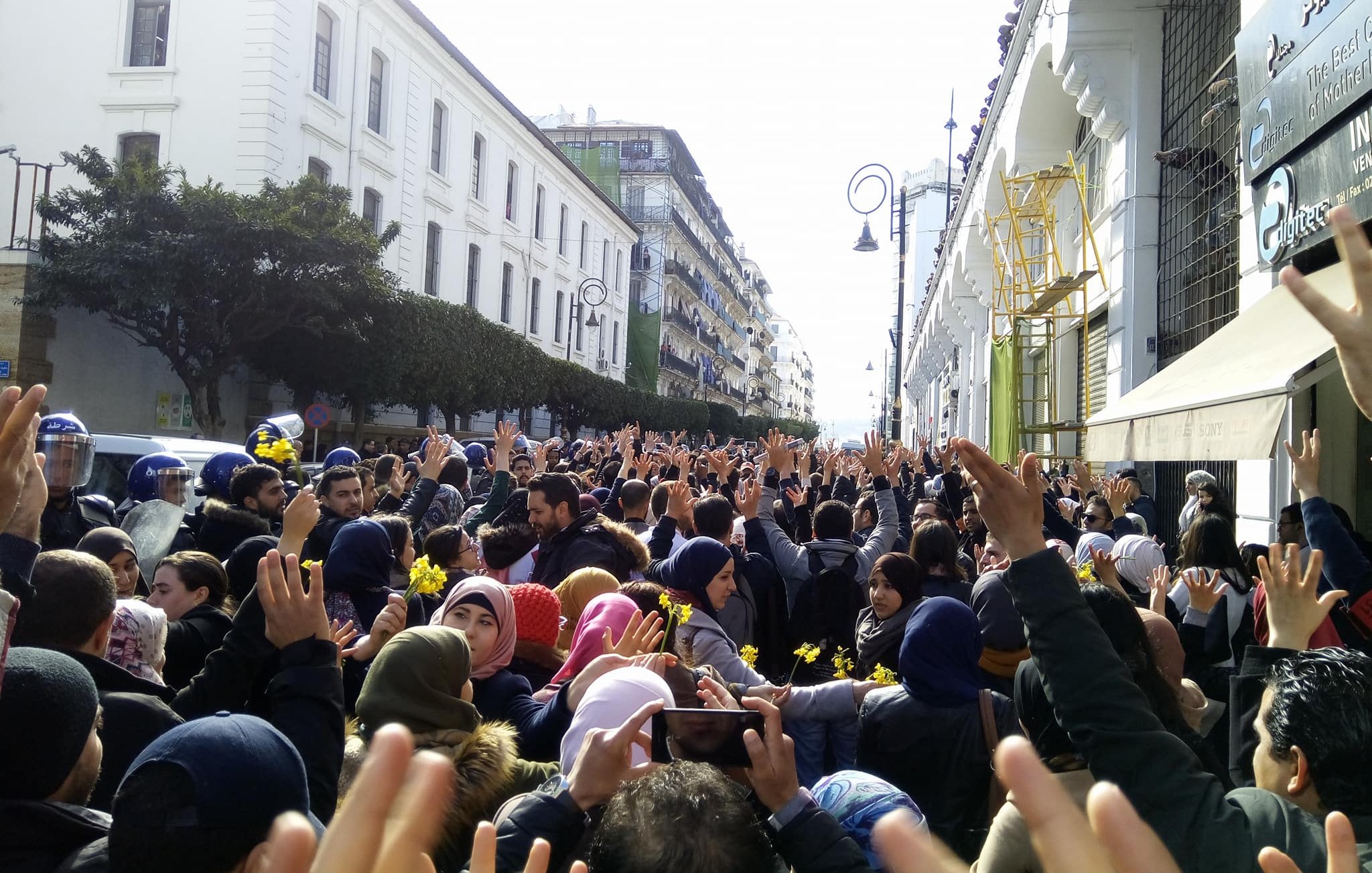 L'image montre une grande foule de personnes rassemblées dans une rue, levant les mains en signe de protestation ou de solidarité. On peut voir des bâtiments architecturaux en arrière-plan, ainsi que quelques agents de police en uniforme. Les participants semblent très engagés dans leur manifestation, et l'atmosphère est chargée d'énergie collective. Les vêtements des manifestants varient, certains portant des écharpes ou des bonnets en fonction de la saison.