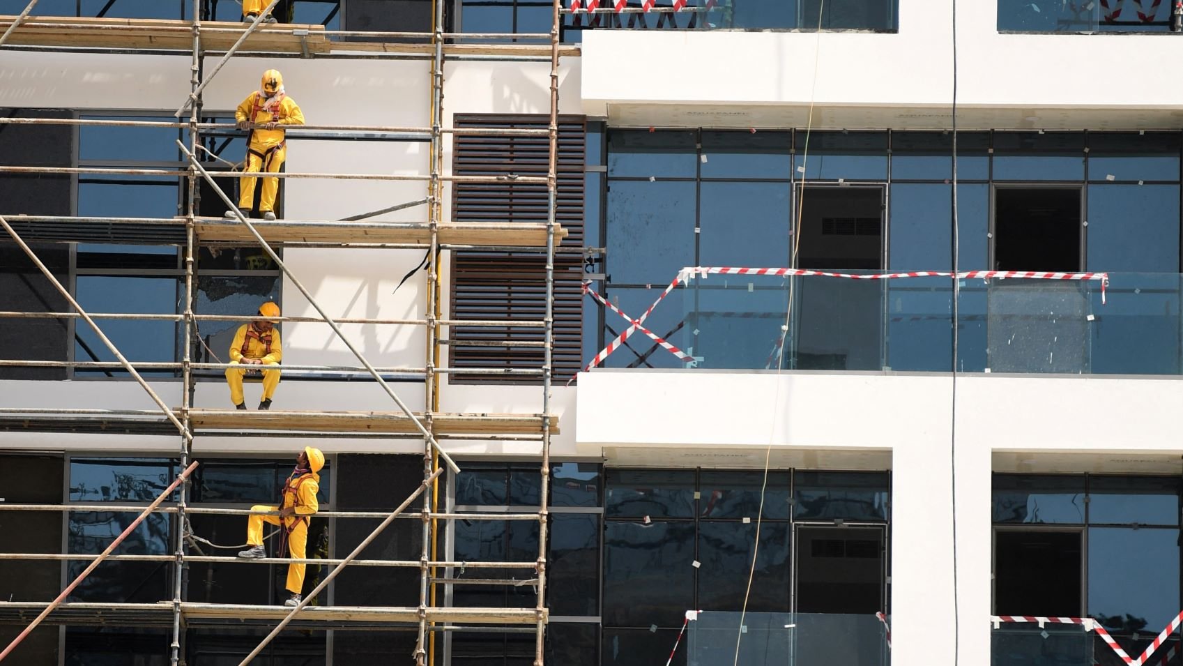 L'image montre un bâtiment en construction. On peut voir des échafaudages le long de la façade, sur lesquels plusieurs ouvriers vêtus de combinaisons jaunes et de casques de sécurité sont en train de travailler. Certains utilisent des outils pour peindre ou poser des éléments sur les fenêtres, tandis que d'autres montent et descendent sur les échafaudages. Le bâtiment a des fenêtres en verre, et des bandes de sécurité sont visibles autour de certaines zones. L'ensemble de la scène reflète un environnement de travail actif et organisé.