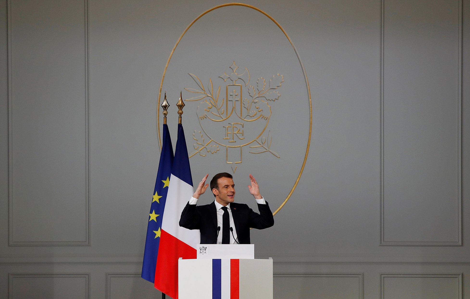 L'image montre un homme debout derrière un pupitre, avec les mains levées comme s'il s'adressait à un auditoire. Derrière lui, il y a un drapeau français et un symbole décoratif sur le mur. Le pupitre présente des couleurs bleu, blanc et rouge, rappelant celles du drapeau français. L'intérieur semble officiel, avec des murs clairs et une ambiance formelle. L'homme semble être en train de faire un discours ou une déclaration importante.