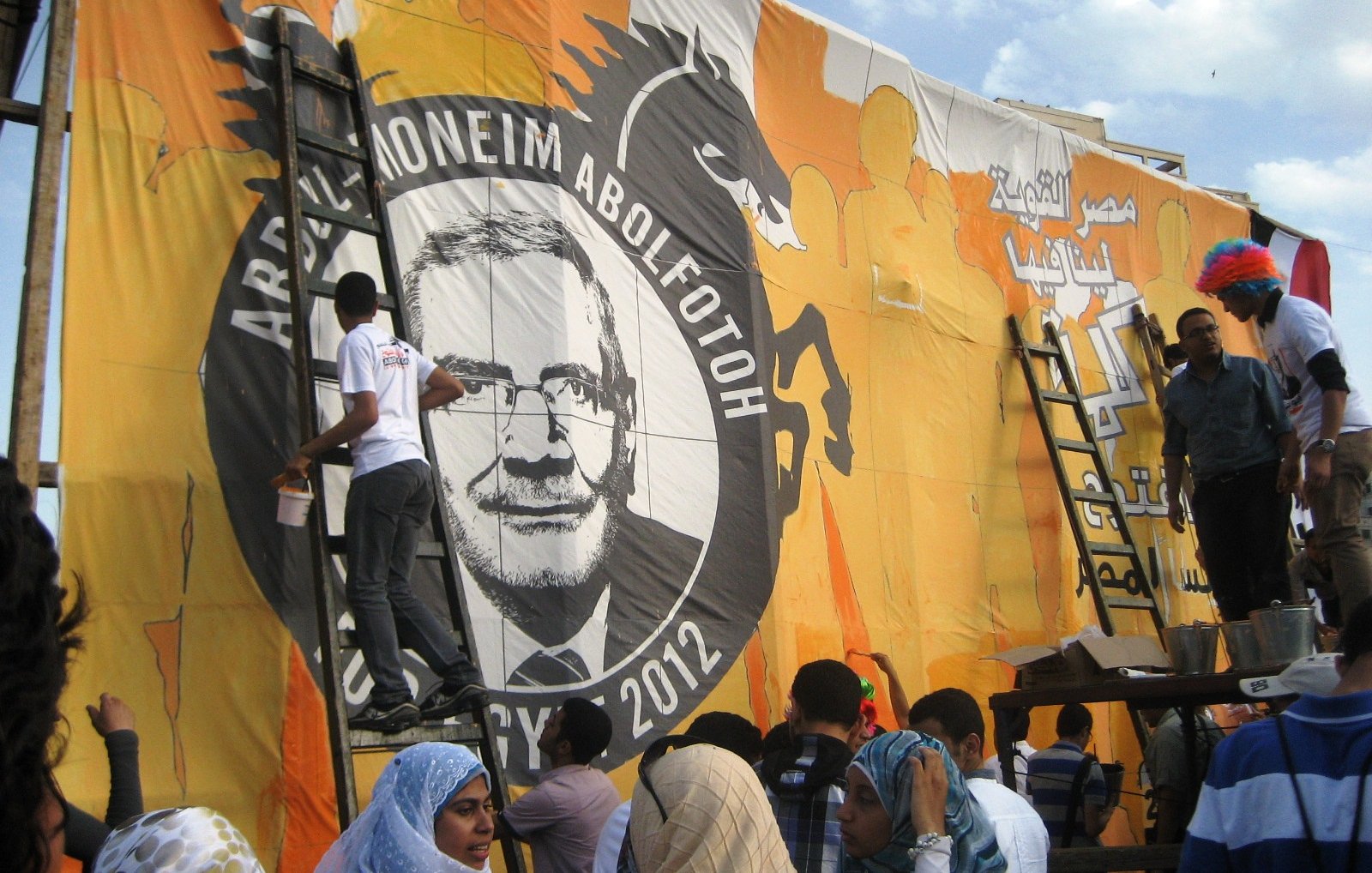 The image shows a group of people working together to paint or prepare a large mural or banner. The mural features a prominent circular design with the name "Abdel-Moneim Abol Fotoh" and the year "2012." There is a dragon motif in the background, which suggests a strong and bold theme. Some individuals are on ladders, actively engaged in applying paint, while others are gathered below, possibly helping or observing. The people are dressed in a variety of clothing styles, and there are some individuals wearing colorful wigs, indicating a festive or celebratory atmosphere. The backdrop is bright orange with various designs.