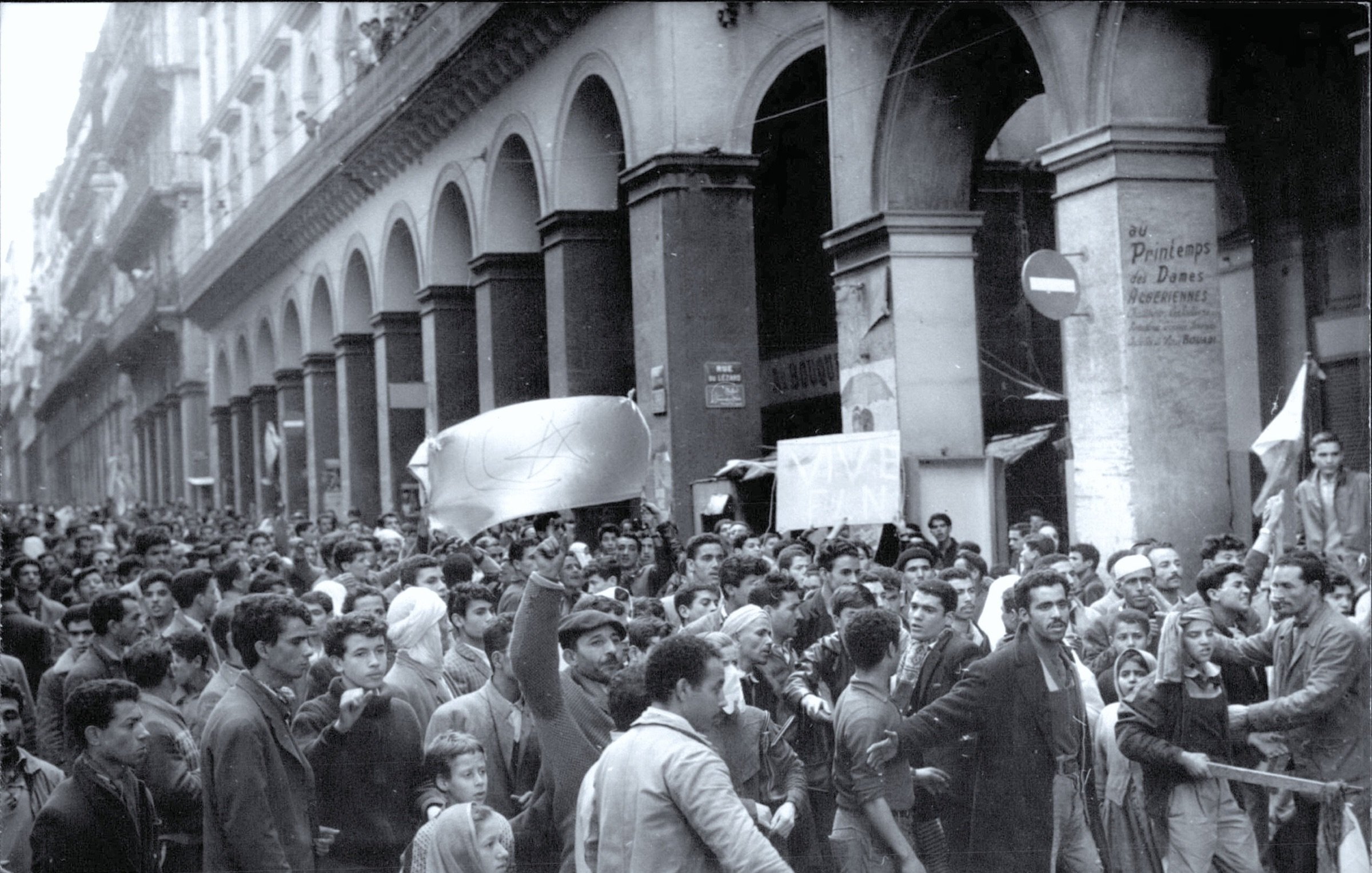 الصورة تظهر تجمعًا حاشدًا لأشخاص يسيرون في أحد الشوارع. الشارع يبدو مزدحمًا بالمتظاهرين، حيث يحمل البعض لافتات وأعلام. يبدو أنهم يعبرون عن مطالب أو احتجاجات. العمارة في الخلفية تعكس طابعًا تاريخيًا وتساهم في خلق جو من الزمان والمكان الذي تتواجد فيه هذه الفعالية. تعبيرات الوجوه تُظهر الحماس والعزيمة بين المشاركين.