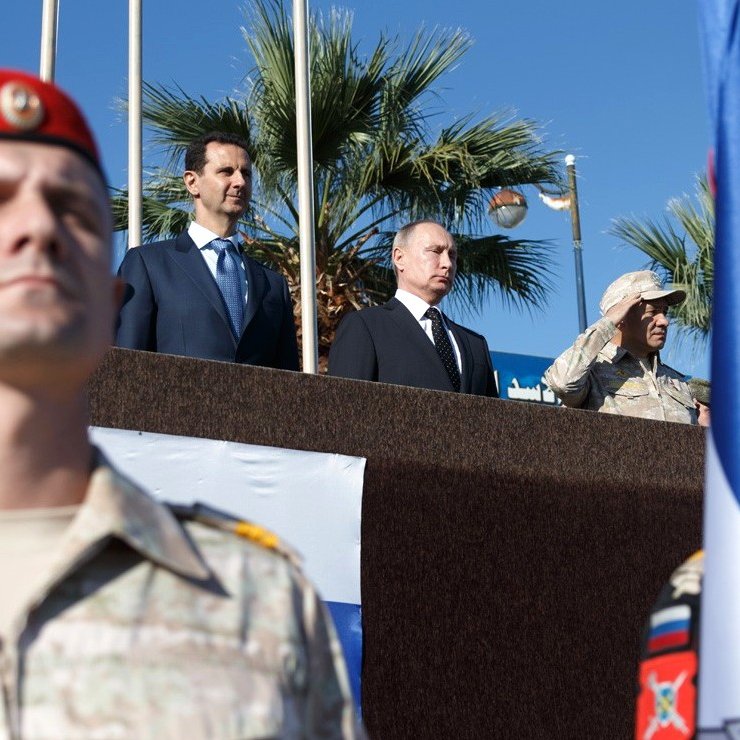 L'image montre un groupe de personnes, dont deux figures de haut rang, sur une plateforme en extérieur, avec un fond de palmiers et un ciel bleu. Au premier plan, des soldats en uniforme, probablement d'une unité militaire, sont présents, portant des drapeaux. Les expressions des personnes sur la plateforme semblent sérieuses et concentrées, indiquant un événement officiel ou militaire.