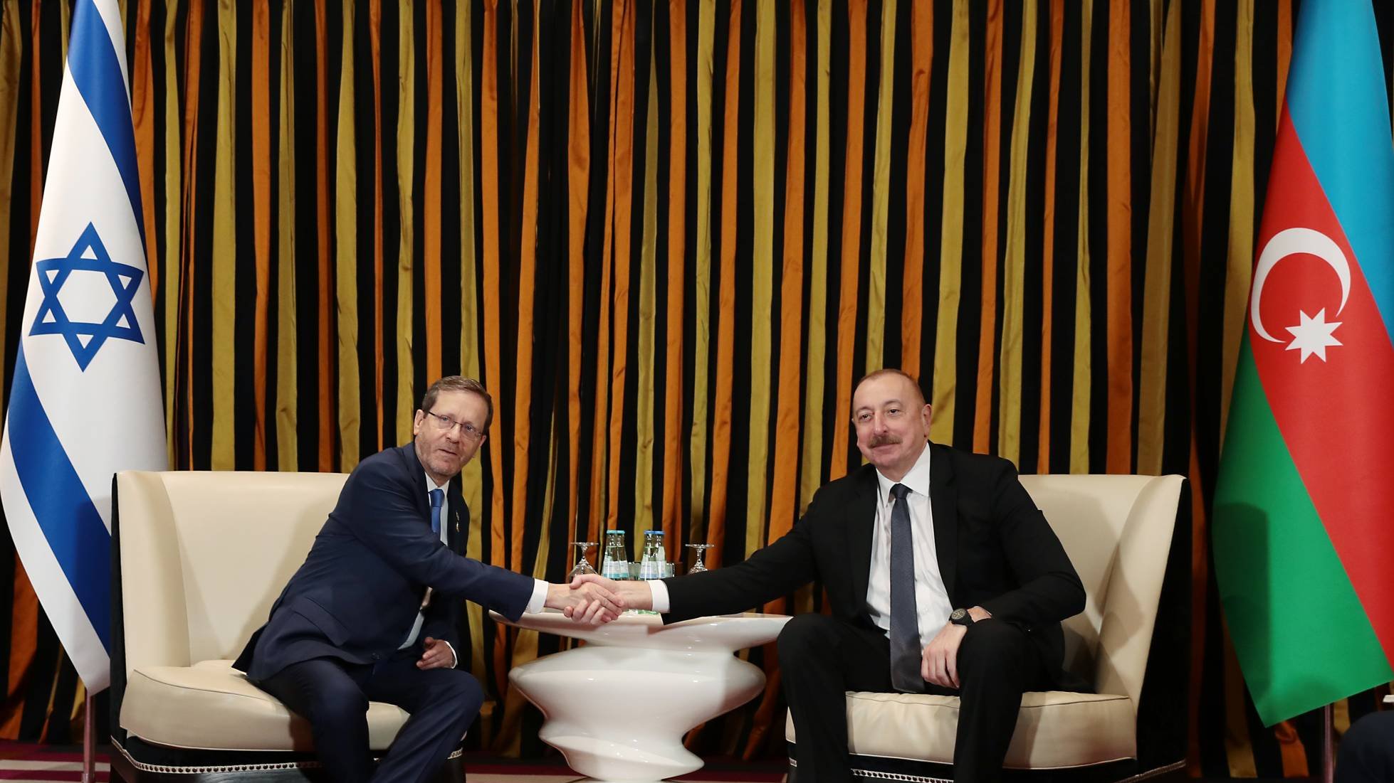 دو مرد در حال دست دادن نشسته‌اند. پرچم‌های اسرائیل و آذربایجان در پس‌زمینه هستند. رنگ‌ها گرم و زنده‌اند.