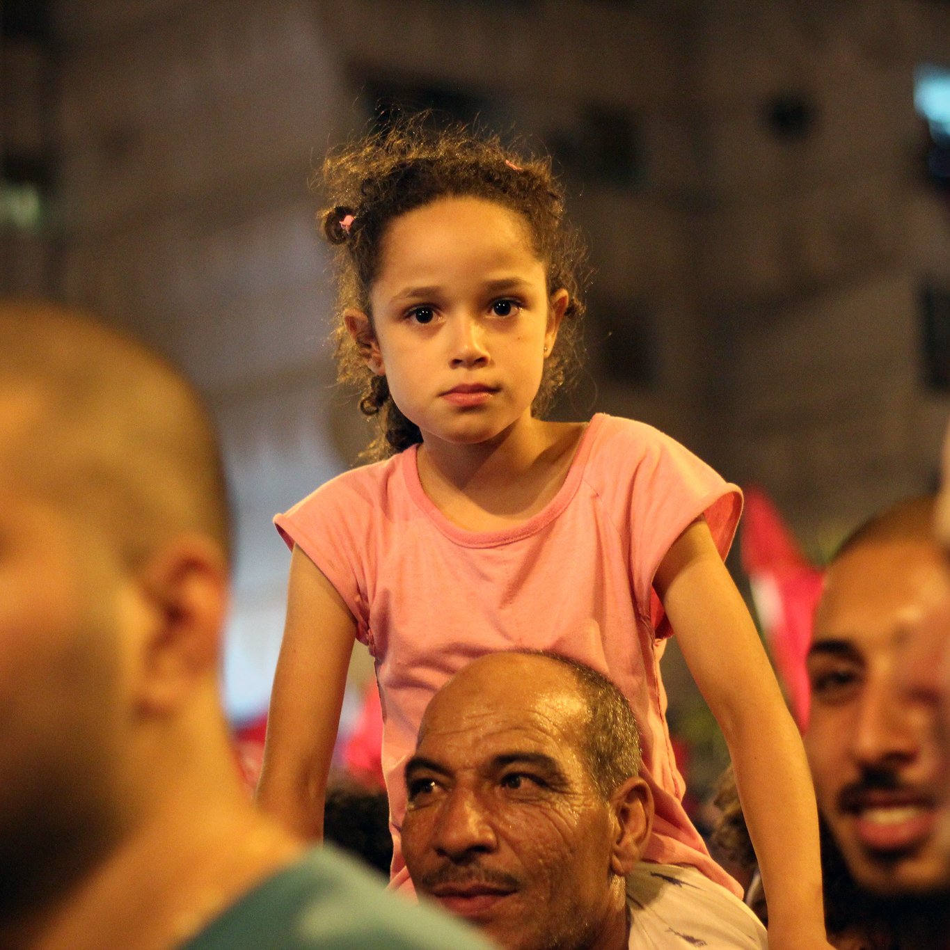 The image depicts a crowd at a public event or gathering. In the foreground, a little girl with curly hair, wearing a pink t-shirt, is seated on the shoulders of a man. She appears thoughtful or concerned, contrasting with the smiling faces of the adults around her. The background suggests a lively atmosphere, possibly filled with people and festive decorations. The scene captures a moment of reflection amid a bustling crowd.