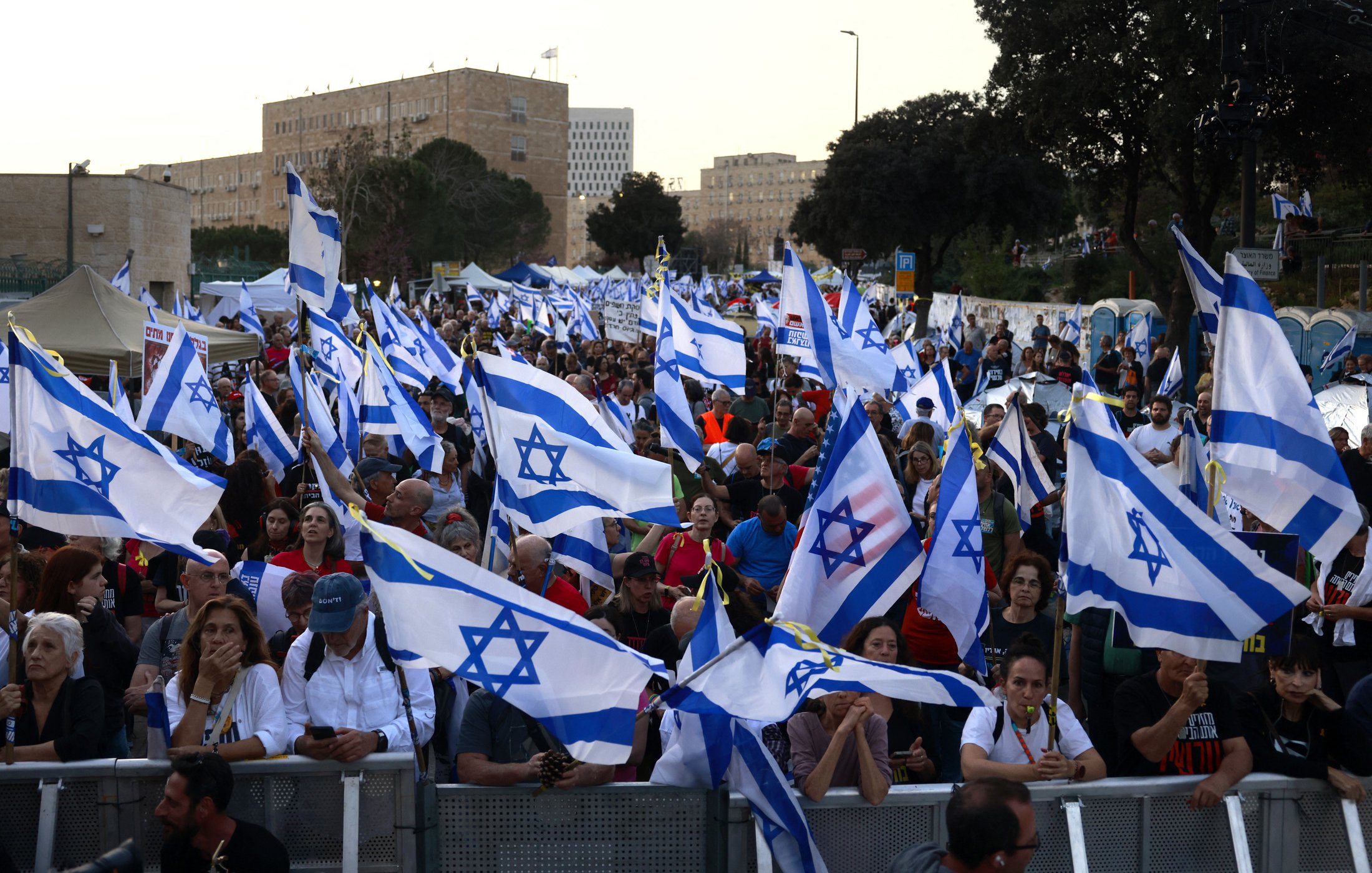 The image shows a large crowd of people gathered together, holding numerous Israeli flags. The atmosphere appears to be part of a demonstration or rally, with many individuals engaged in conversation or focused on the event. The background features buildings and trees, indicating an urban setting. The flags are prominently displayed, creating a sense of unity among the participants.