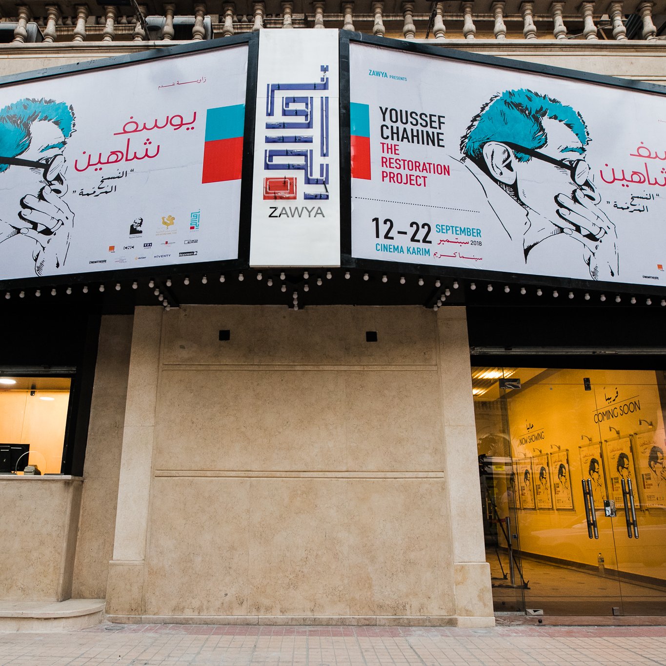 L'image montre un bâtiment avec des bannières affichant un événement intitulé "Youssef Chahed - The Restoration Project". Les bannières présentent un portrait stylisé, probablement d'un homme portant un masque. La date de l'événement est indiquée, s'étalant du 12 au 22 septembre. On remarque également une ambiance artistique et contemporaine, suggérant que cet événement est lié à l'art ou à une exposition. L'entrée du bâtiment semble accueillante, avec un guichet visible.