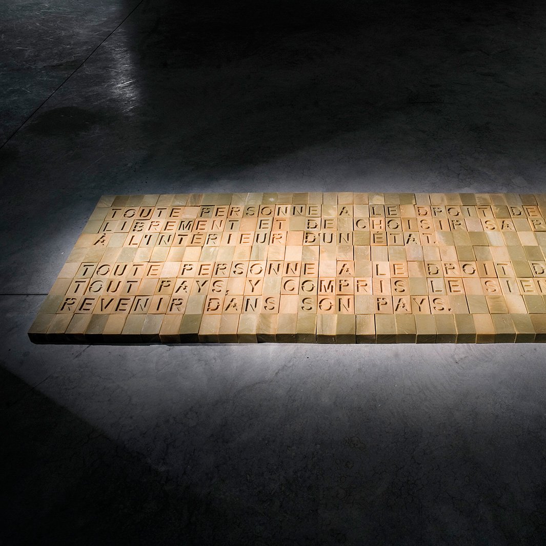 L'image montre une installation au sol formée de blocs rectangulaires disposés de manière à former un texte. Le texte est en relief et semble évoquer des thèmes liés aux droits de l'homme, notamment le droit à la liberté et à l'asile. L'éclairage met en valeur les mots, créant une ambiance à la fois réfléchie et sérieuse. Le sol sur lequel se trouvent les blocs est en béton, et l'environnement semble être une galerie d'art ou un espace d'exposition.