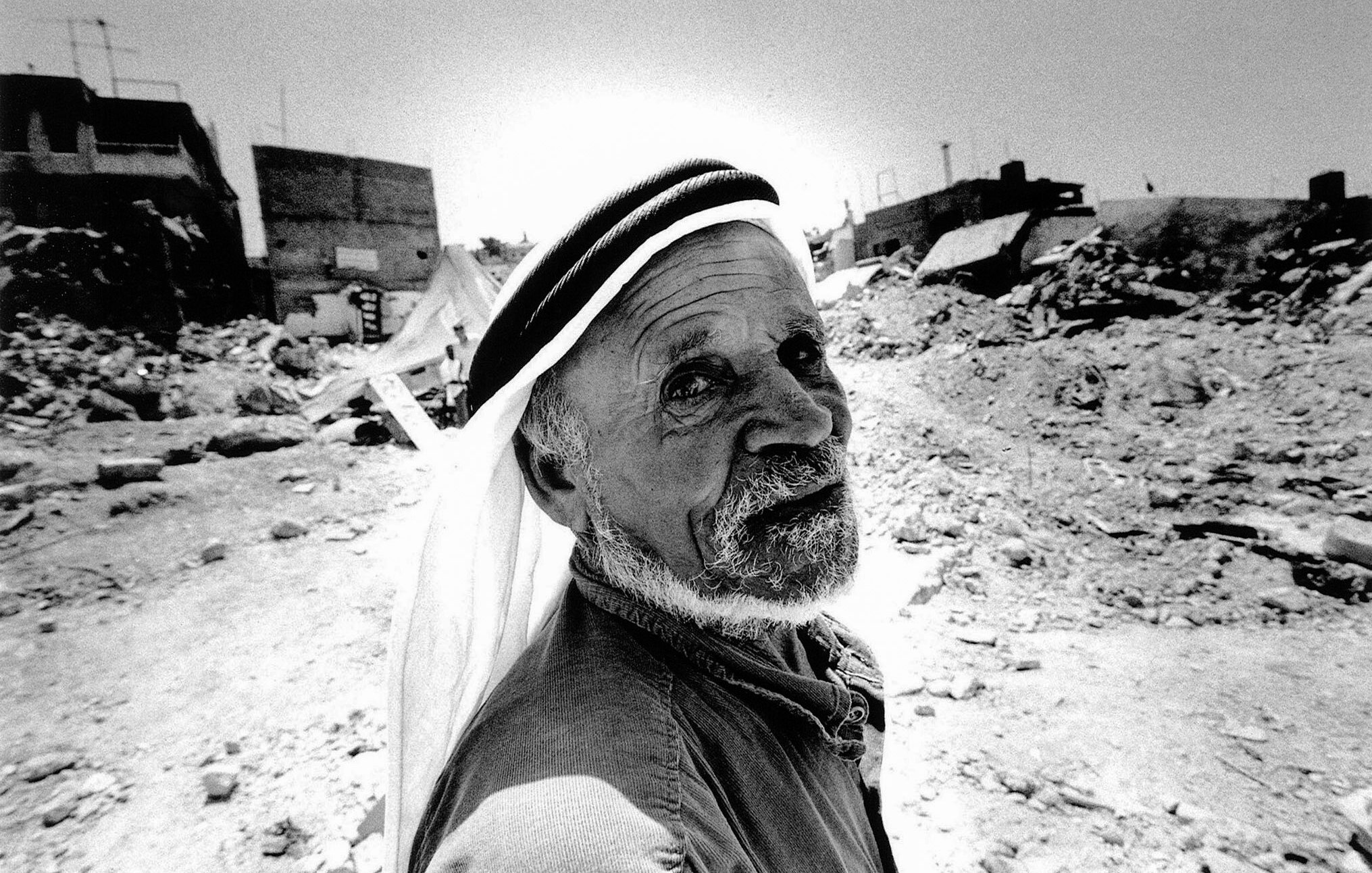 L'image montre un homme âgé au visage marqué, qui regarde directement vers l'objectif. Il porte un keffieh, un couvre-chef traditionnel, et a une expression qui dégage une certaine sagesse ou tristesse. En arrière-plan, on peut apercevoir des ruines, des débris et des bâtiments partiellement détruits, suggérant un environnement difficile ou une situation de conflit. La scène est capturée en noir et blanc, ce qui accentue les textures et les contrastes des éléments environnants.
