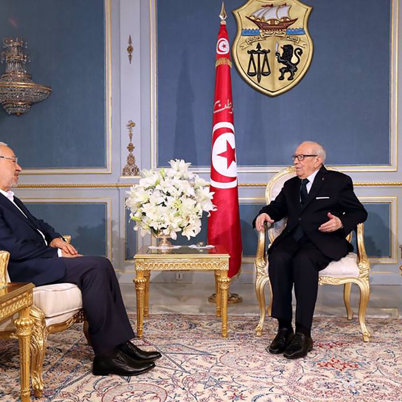 L'image montre deux hommes assis dans un cadre officiel, probablement dans un bureau ou une salle de réunion élégante. Les deux sont vêtus de costumes sombres, l'un portant des lunettes. Au fond, on peut voir le drapeau tunisien ainsi qu'un blason. La pièce est décorée avec des meubles en or, des fleurs blanches et un tapis coloré, ce qui crée une atmosphère formelle et importante.