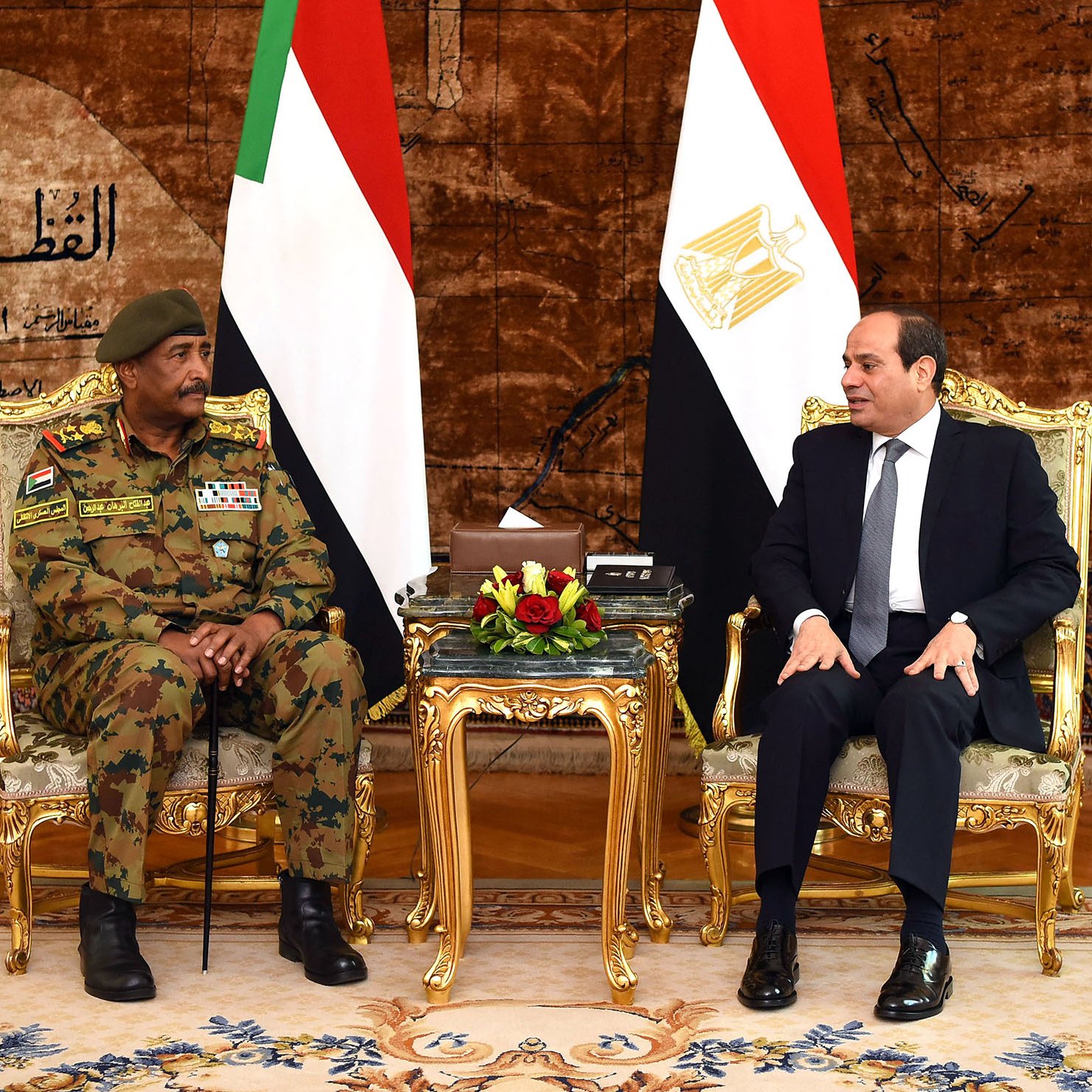 تظهر الصورة لقاءً رسميًا بين شخصين يجلسان على كراسي فاخرة أمام علمي السودان ومصر. خلفهم يوجد ستار مزخرف يضيف لمسة فخمة للمكان. الشخص على اليسار يرتدي زيًا عسكريًا ويبدو أنه مسؤول عسكري، بينما الشخص على اليمين يرتدي بدلة رسمية. يعبر كلا الشخصين عن تواصل دبلوماسي بين البلدين.