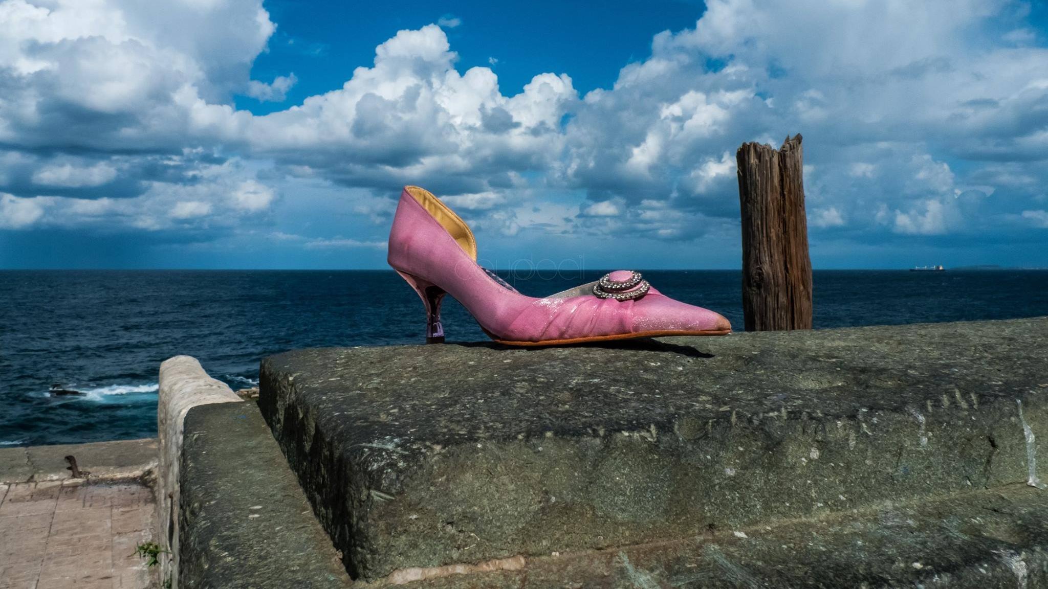 L'image montre une chaussure à talon rose, posée sur un mur en pierre. En arrière-plan, on peut voir une mer calme sous un ciel rempli de nuages blancs et d'un bleu éclatant. À droite, un poteau en bois émerge de la surface, ajoutant une touche de rusticité au paysage. L'ensemble évoque un mélange de solitude et de beauté naturelle.