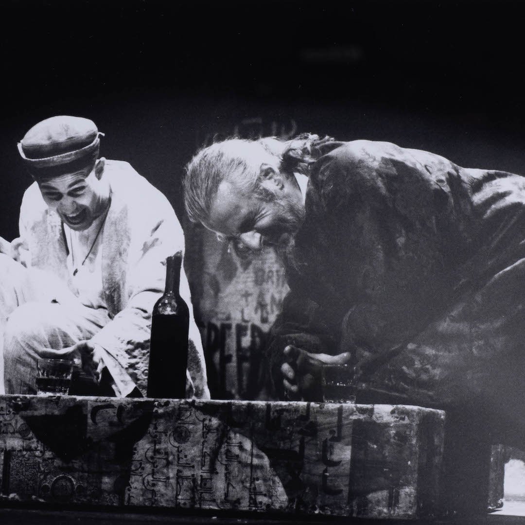 Deux hommes en scène, riant, près d'une bouteille sur une caisse au fond sombre.