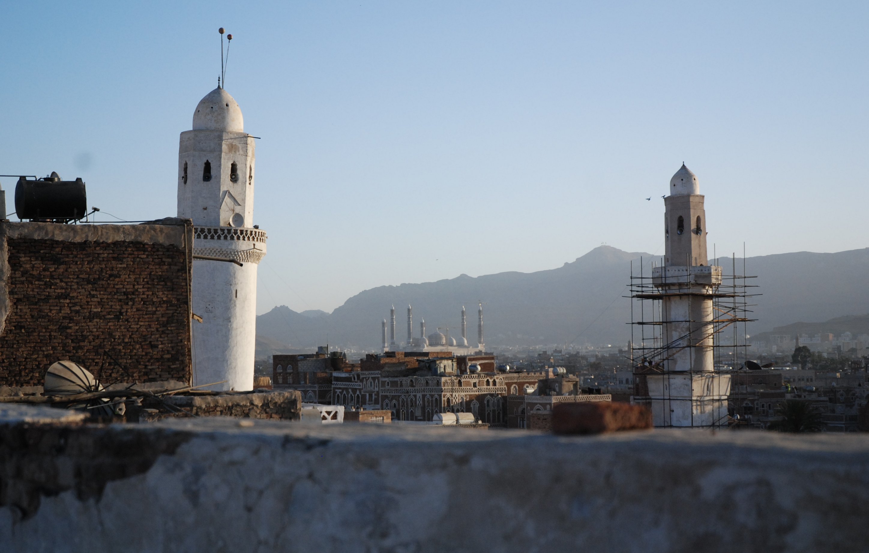 L'image montre une vue panoramique d'une ville avec des minarets distinctifs. On peut voir deux tours blanches caractéristiques, l'une étant en cours de restauration, entourées d'un paysage montagneux en arrière-plan. Le ciel est clair, indiquant une ambiance paisible au crépuscule. Au loin, plusieurs autres structures architecturales, probablement des mosquées, se dessinent à l'horizon, contribuant à l'atmosphère culturelle et historique de la scène.