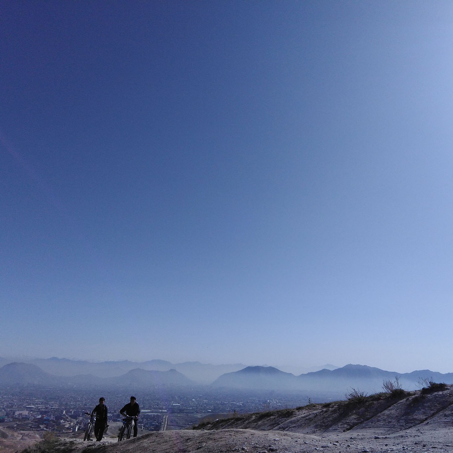 L'image montre deux personnes se tenant sur une colline, contemplant un vaste paysage sous un ciel bleu clair. On peut voir des montagnes à l'horizon et une ville en contrebas, légèrement obscurcie par une brume. La lumière est douce, créant une ambiance paisible et tranquille. Les silhouettes des personnes ajoutent une note humaine à cette scène naturelle.