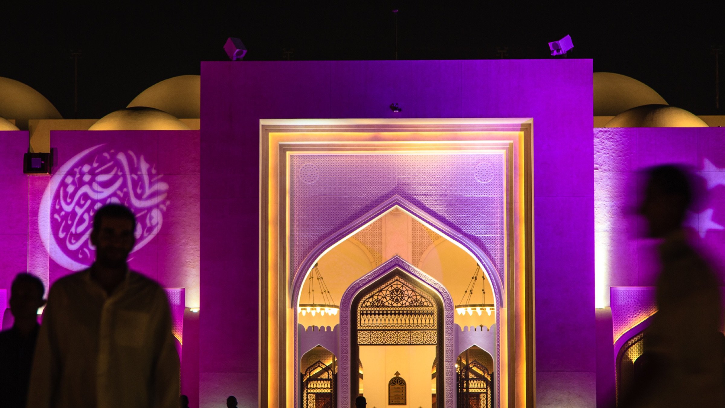 L'image montre une grande structure architecturale éclairée de manière colorée, principalement en violet. On peut voir un portail monumental avec des motifs décoratifs, typiques de l'architecture islamique. À l'avant, des silhouettes de personnes marchent, créant une atmosphère vivante et dynamique. Au fond, des éléments de design comme des arcs et des motifs sont visibles, ajoutant une touche d'élégance à l'ensemble de la scène. L'éclairage nocturne met en valeur les détails architecturaux et crée une ambiance fascinante.