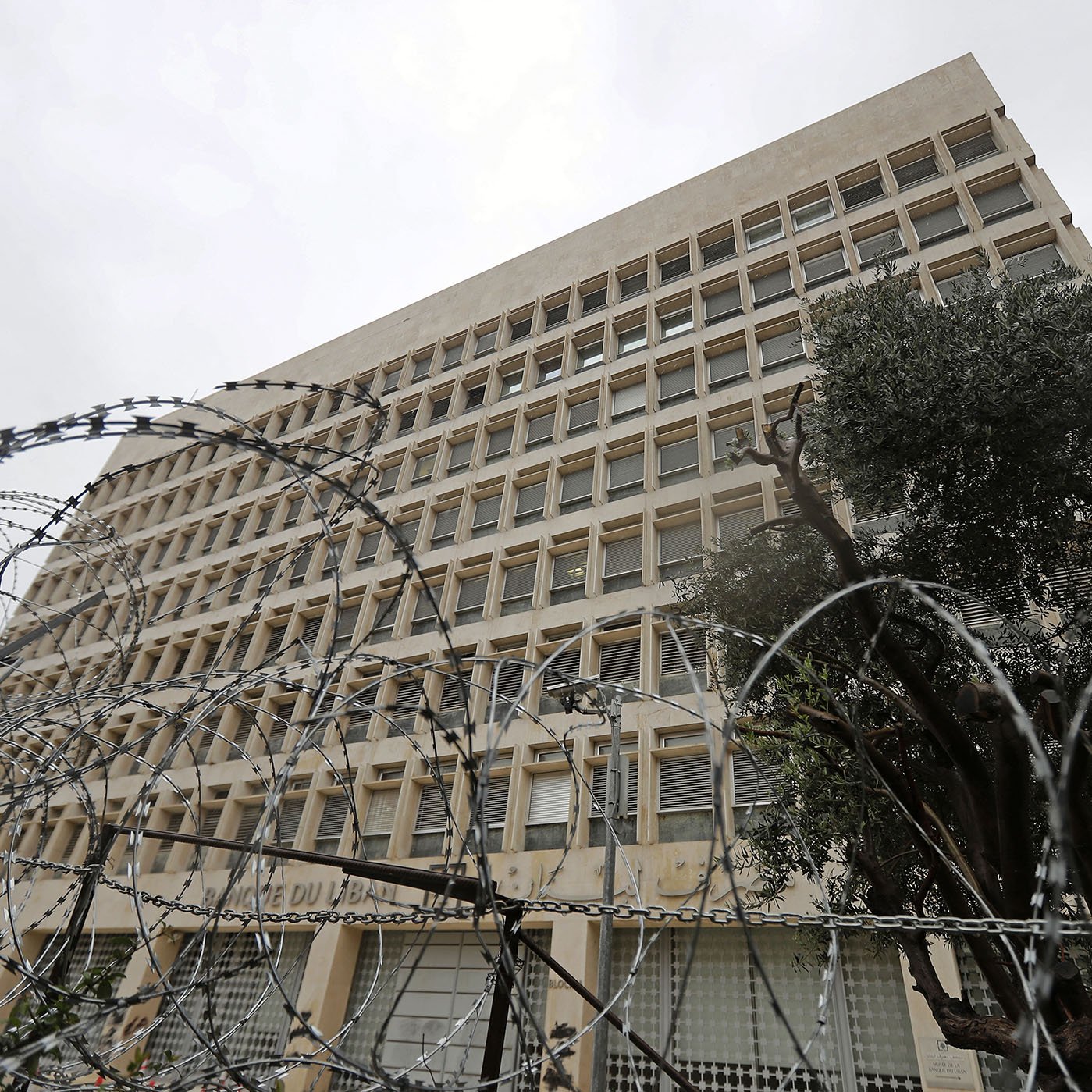 The image shows a large, modern building with a distinct architectural style, featuring multiple windows arranged in a grid pattern. In the foreground, there is a fence topped with barbed wire, indicating a level of security. The scene appears cloudy or overcast, giving a somewhat somber atmosphere. Additionally, there are some trees visible, possibly to the side of the building, adding a touch of greenery to the environment.
